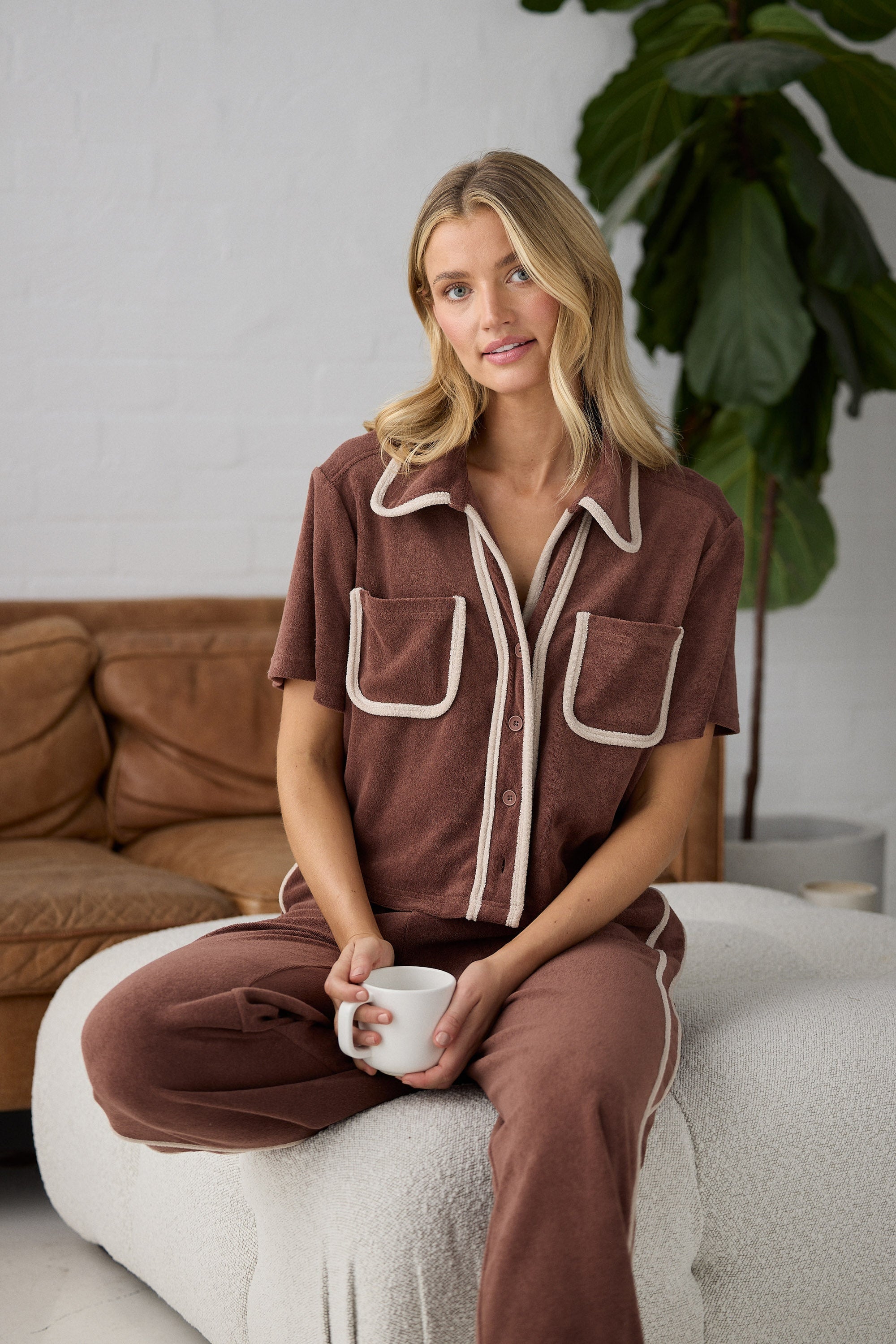 Woman in brown set holding a mug, sitting on a white chair with a plant in the background.