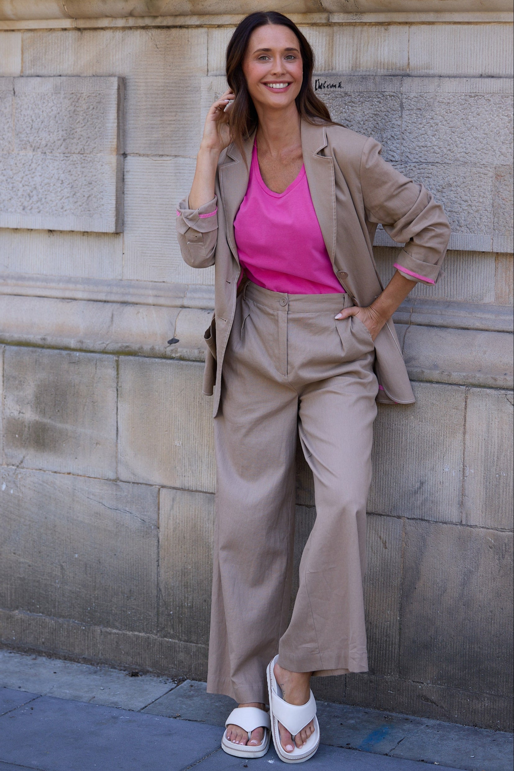 Woman in a beige suit and pink top standing against a stone wall.