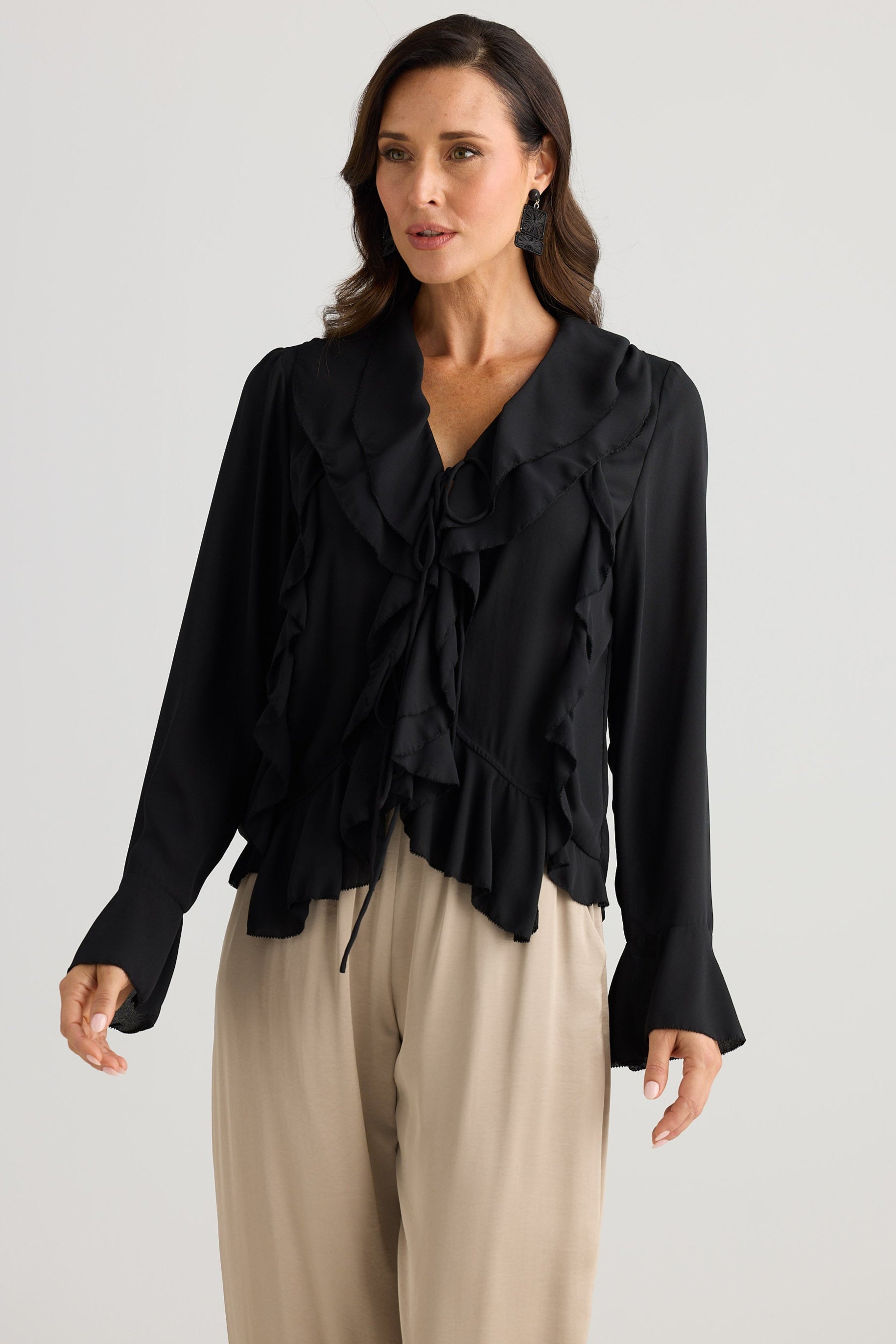 Woman wearing a black blouse with ruffled details and beige pants on a white background