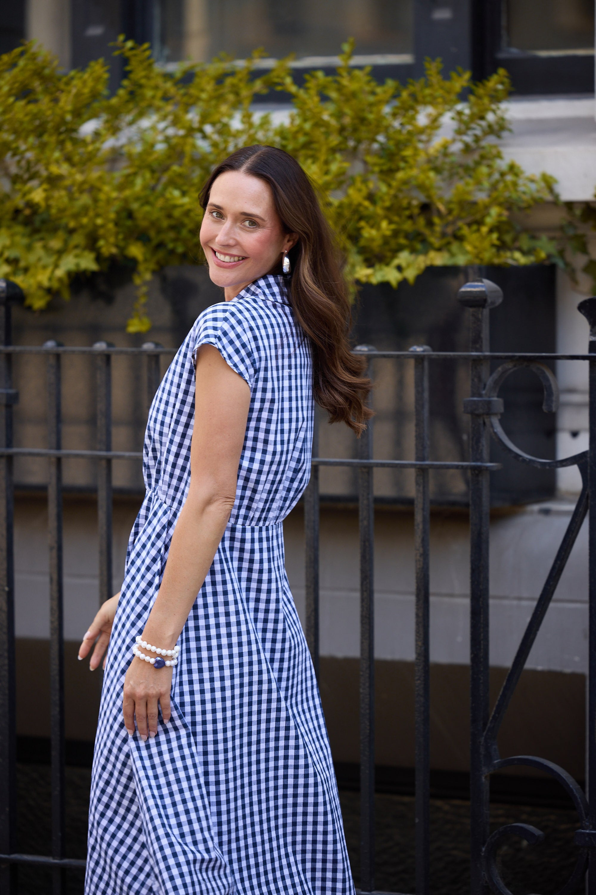Woman in a blue checkered dress standing outdoors with greenery in the background