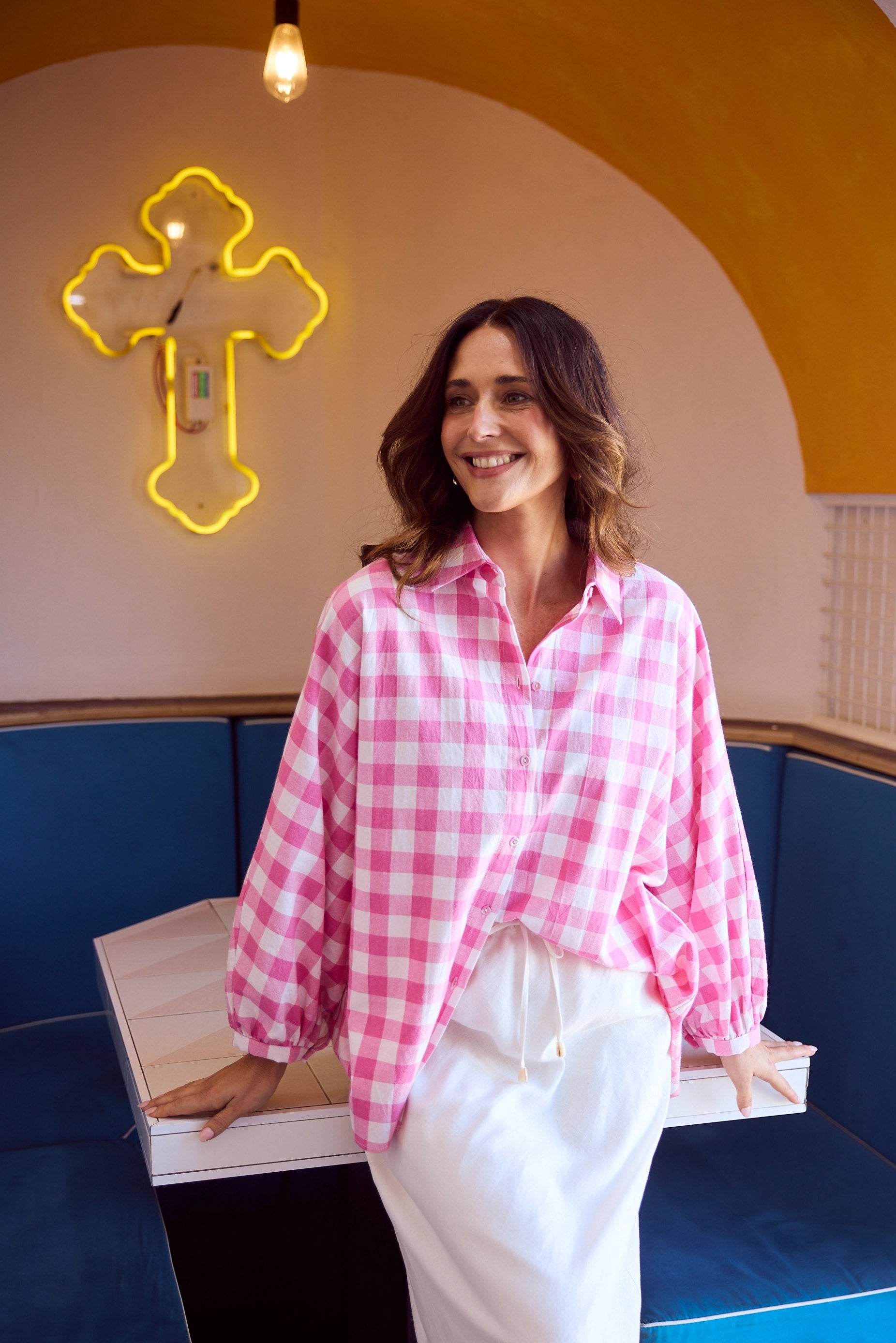 Woman in a pink checkered shirt and white pants standing in a room with blue benches and a yellow neon sign.