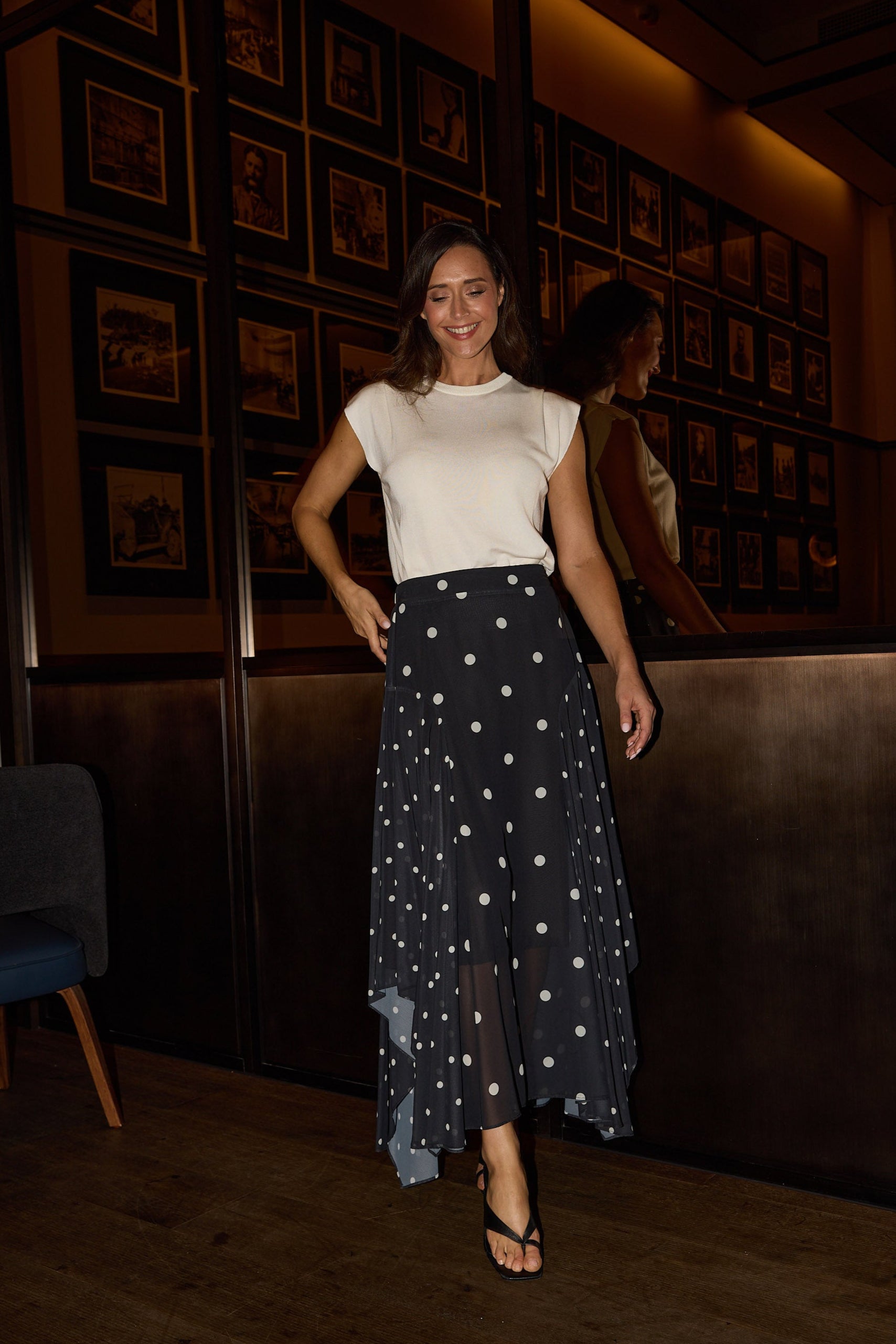 Woman in a white top and dark patterned skirt standing in a dimly lit room with framed pictures on the wall.