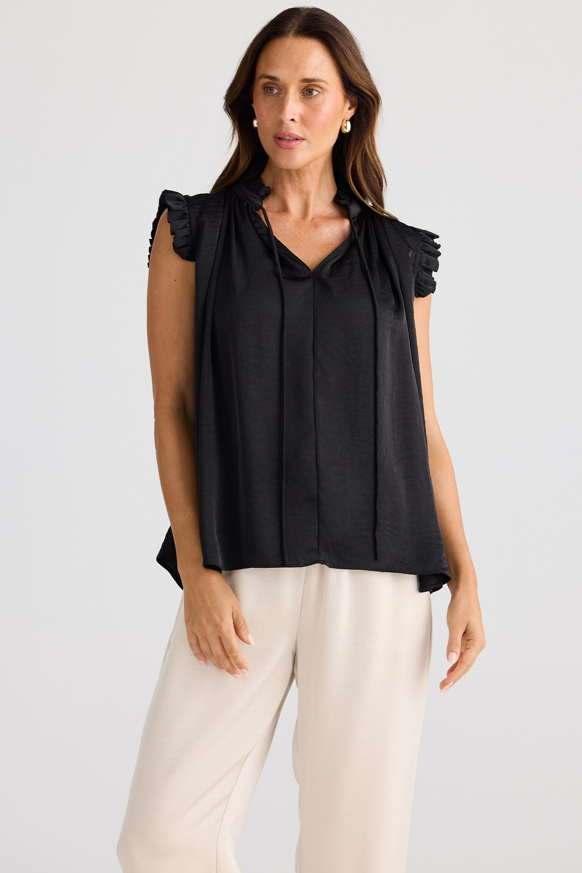 Woman wearing a black blouse with ruffled shoulders and white pants on a light gray background