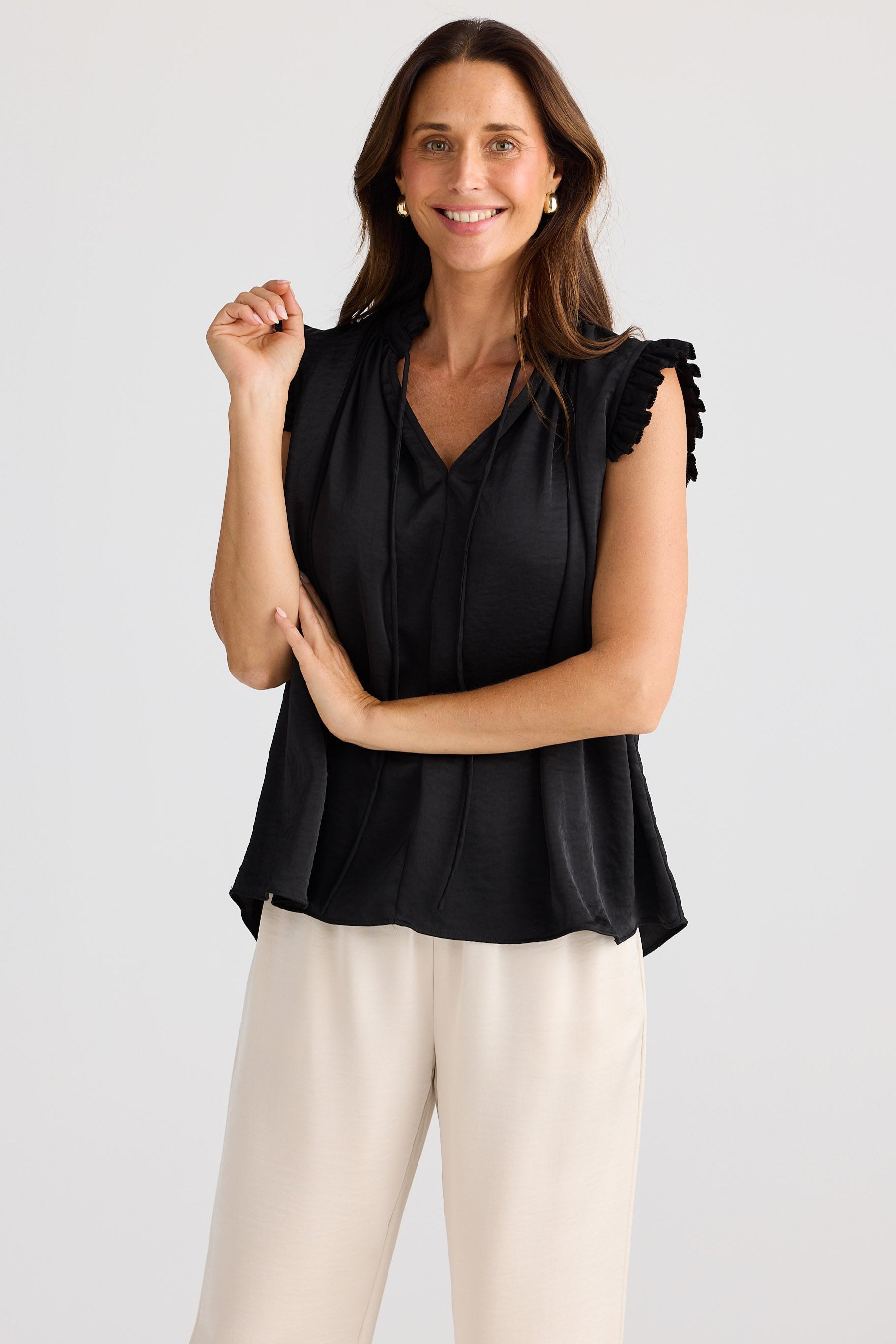 Woman wearing a black sleeveless top with ruffled shoulders and beige pants on a white background