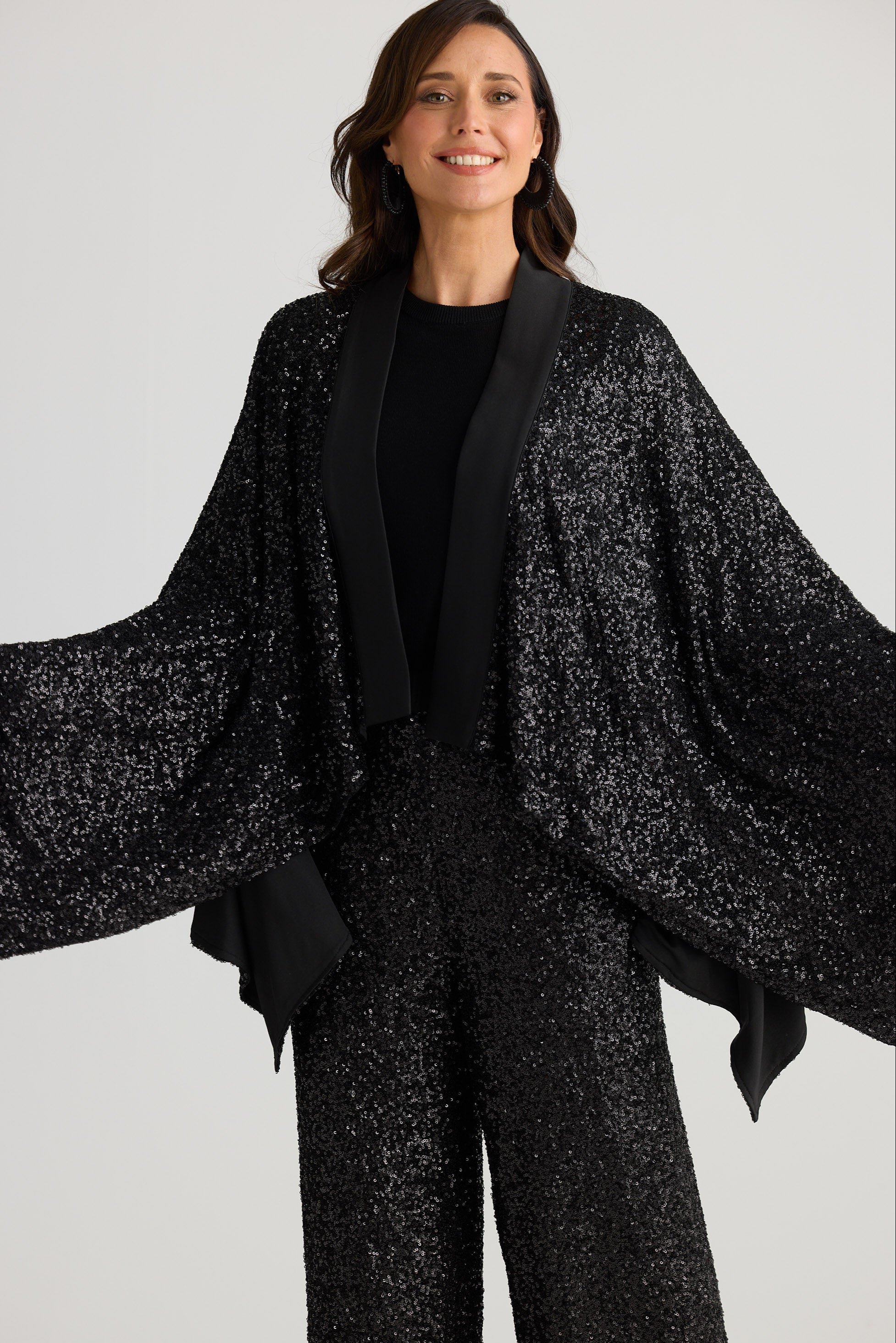 Woman wearing a black textured outfit with a cape-like garment on a light gray background
