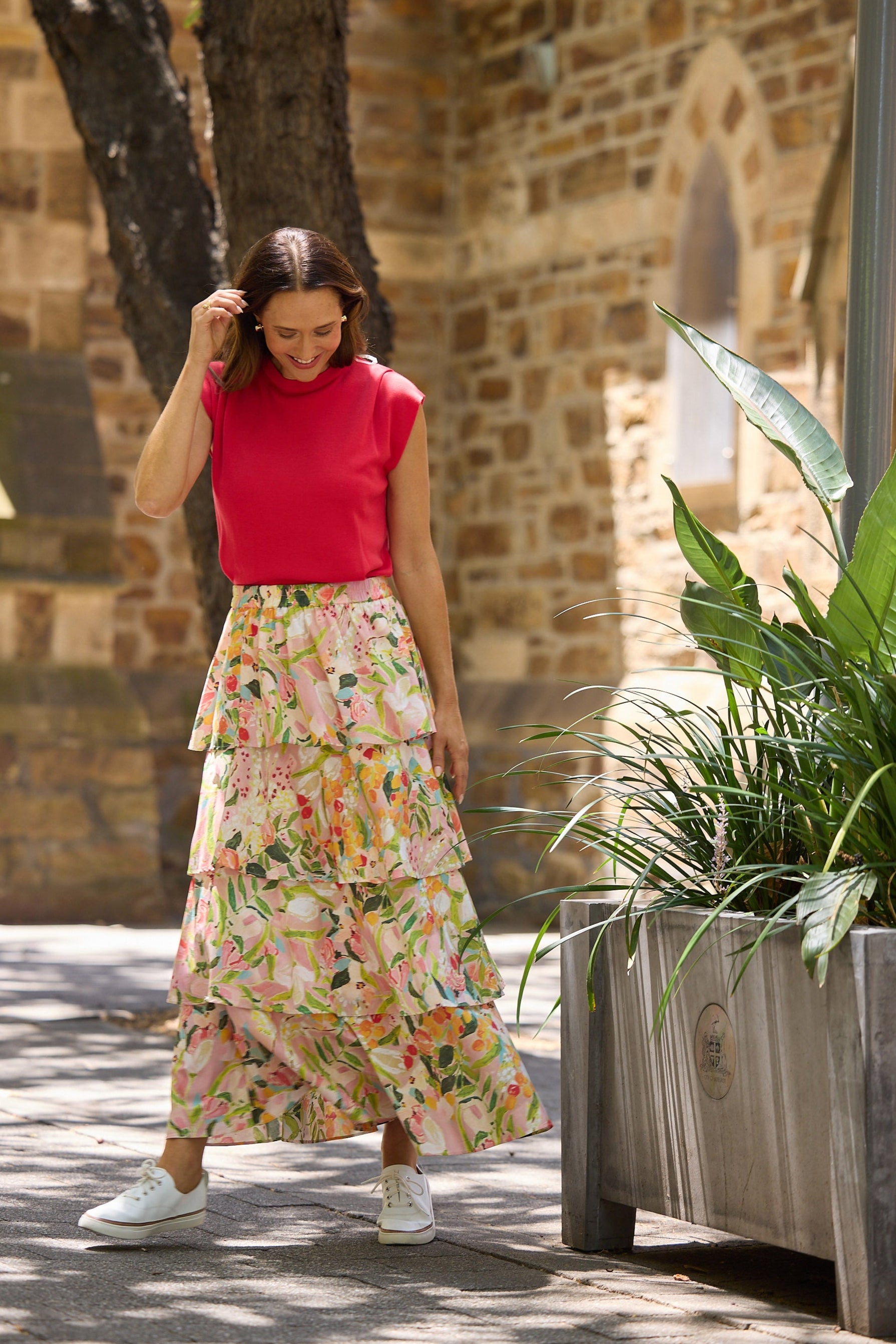 Woman in a red top and floral skirt walking outdoors near a stone building and plants.