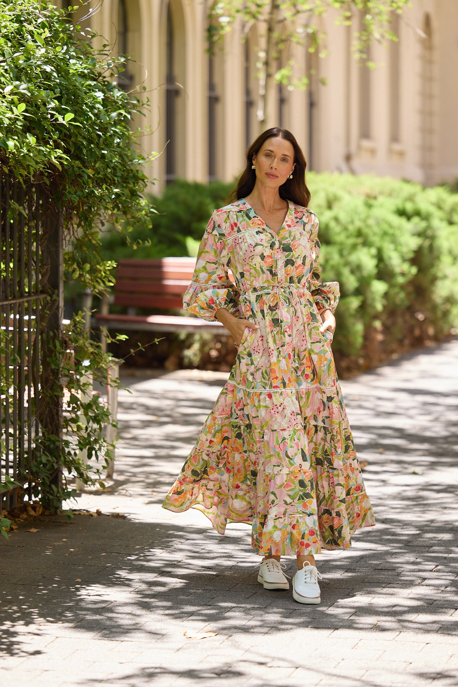 Woman in a floral dress walking on a sunlit path with greenery and a building in the background
