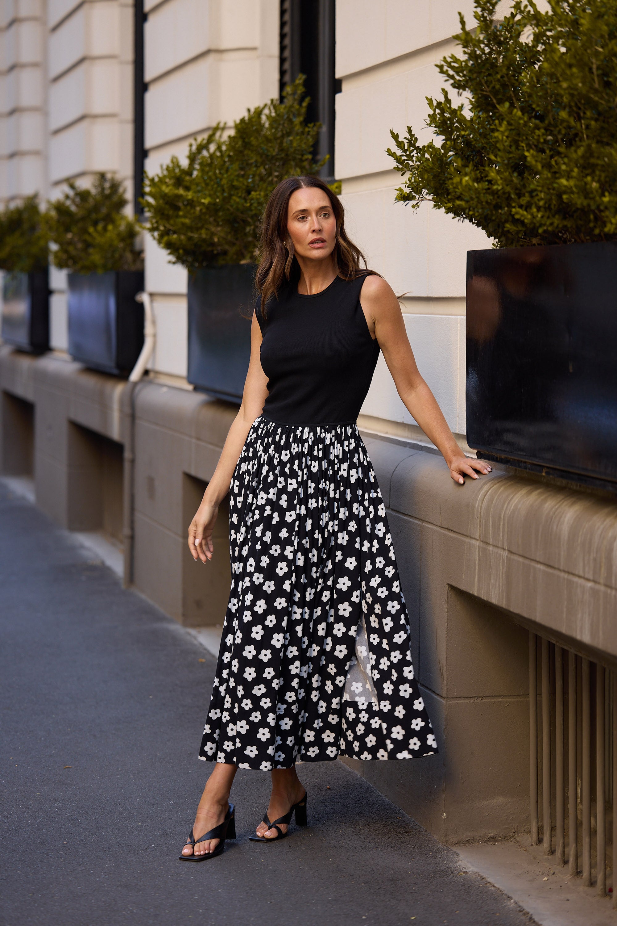 Woman in a dress with black sleeveless top and floral skirt standing on a sidewalk with plants in the background.