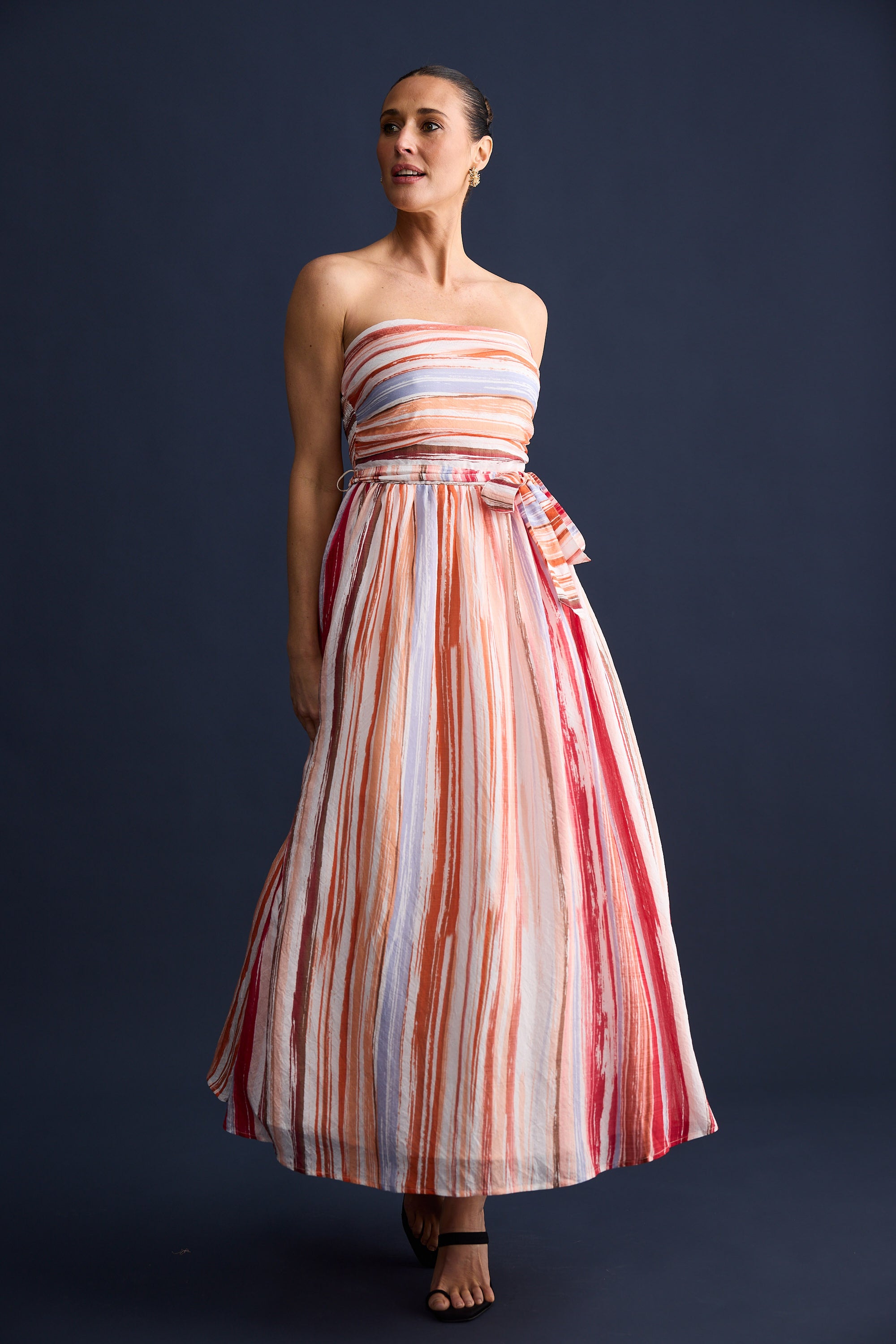 Woman wearing a strapless, long, multicolored striped dress against a dark background