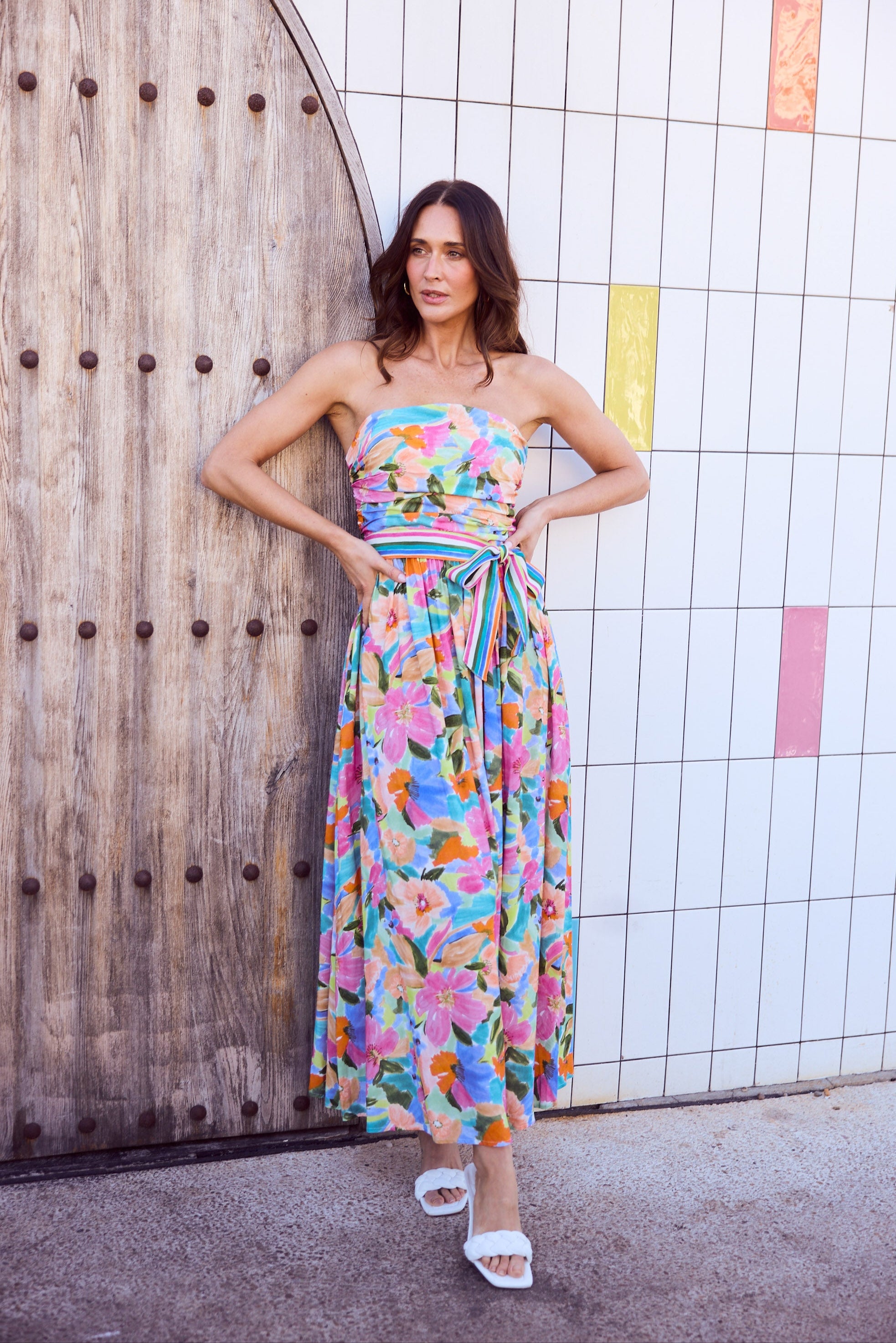 Woman wearing a colorful floral dress standing against a tiled wall.