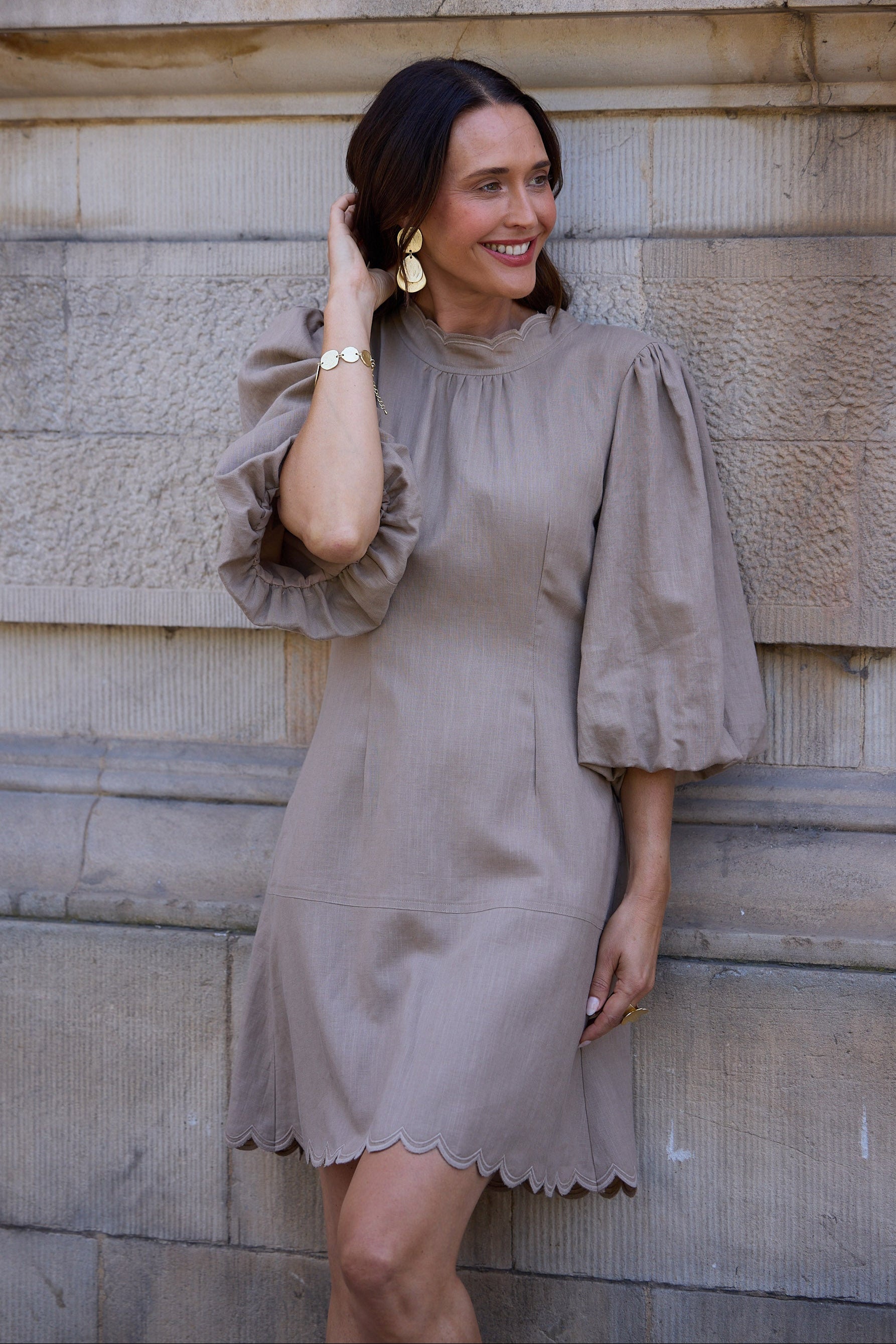 Woman wearing a beige dress with ruffled sleeves against a stone wall.