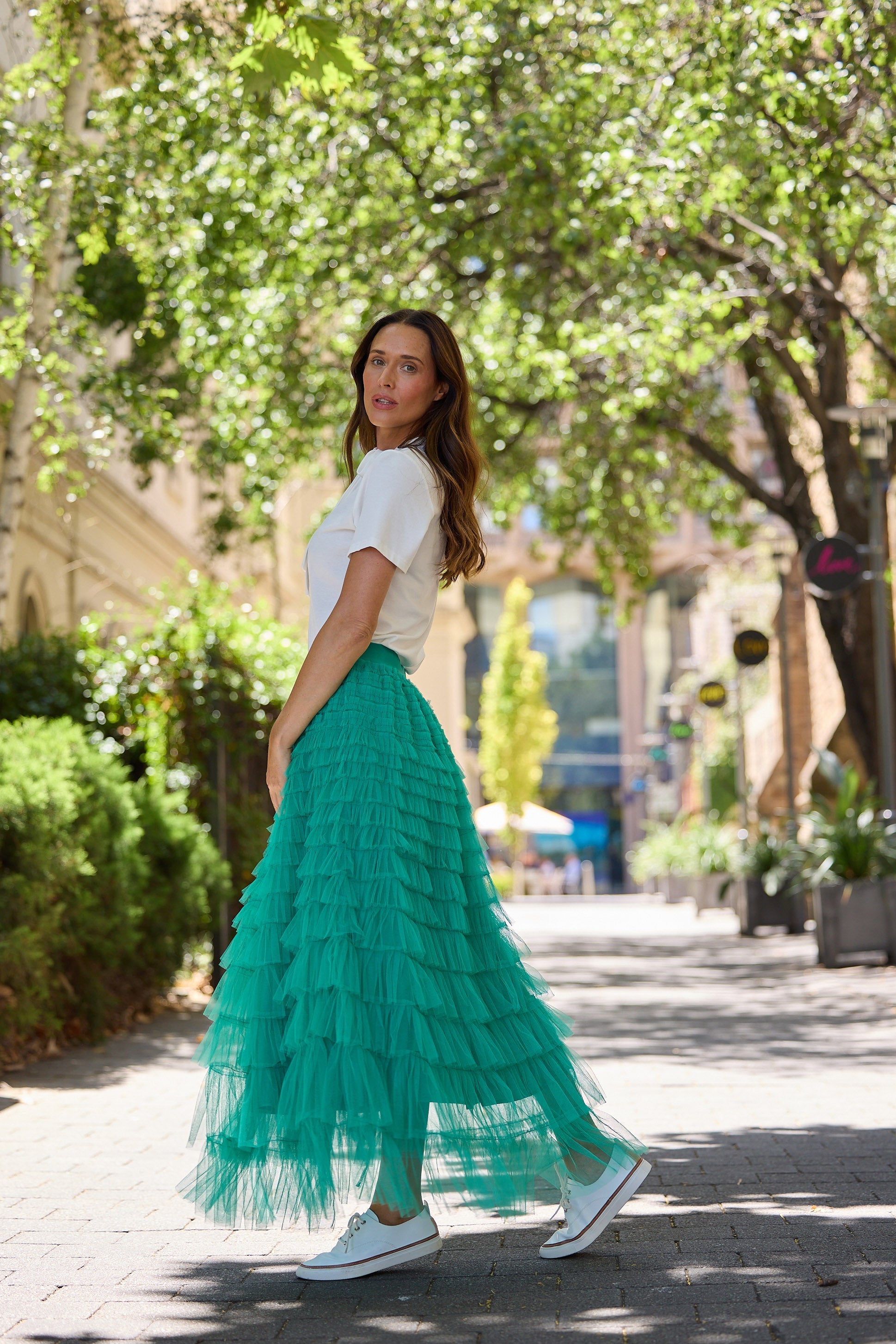 Woman in a white top and teal skirt walking on a street with trees and buildings in the background.