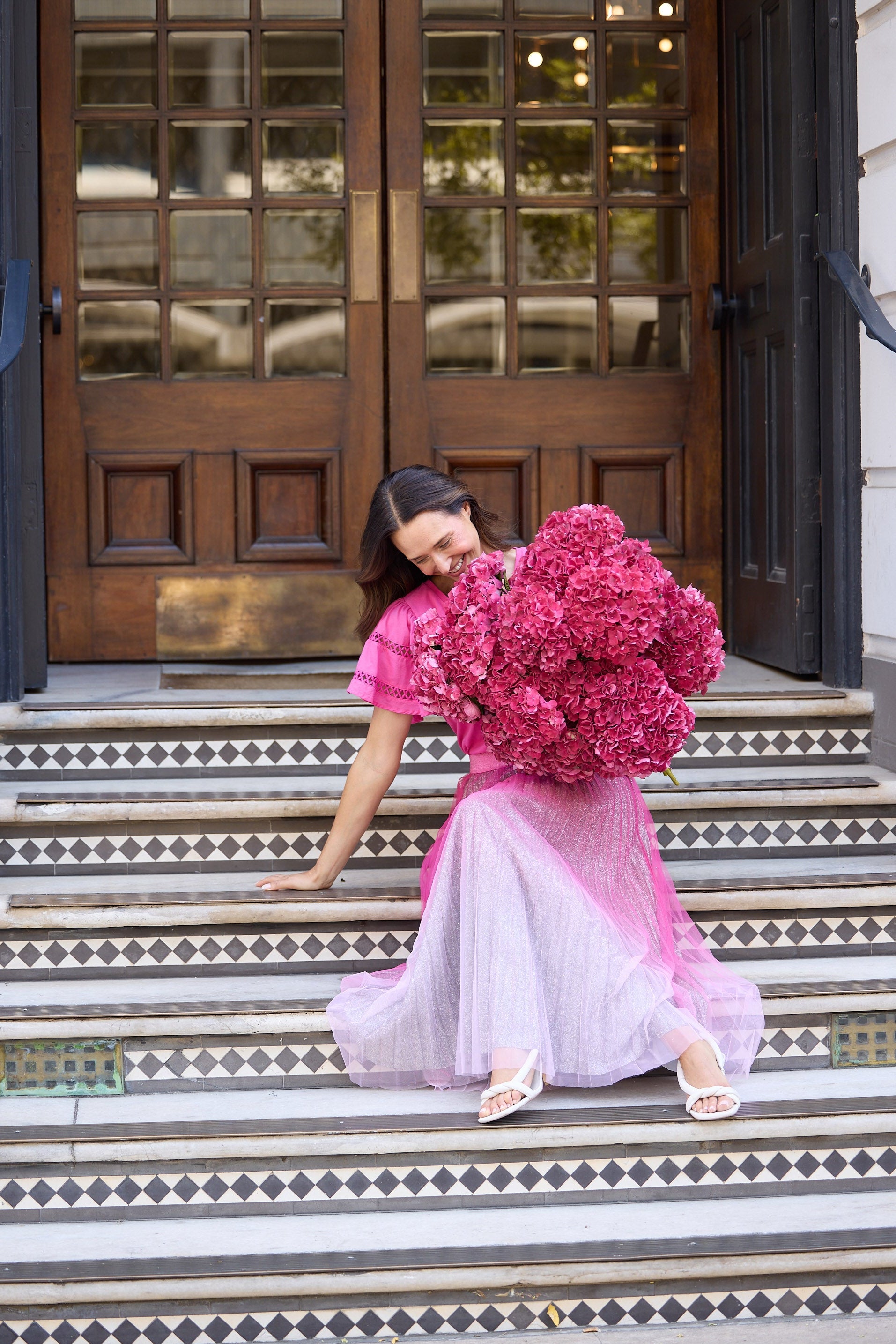 Woman in a pink dress holding a large bouquet of pink flowers on steps outside a building.
