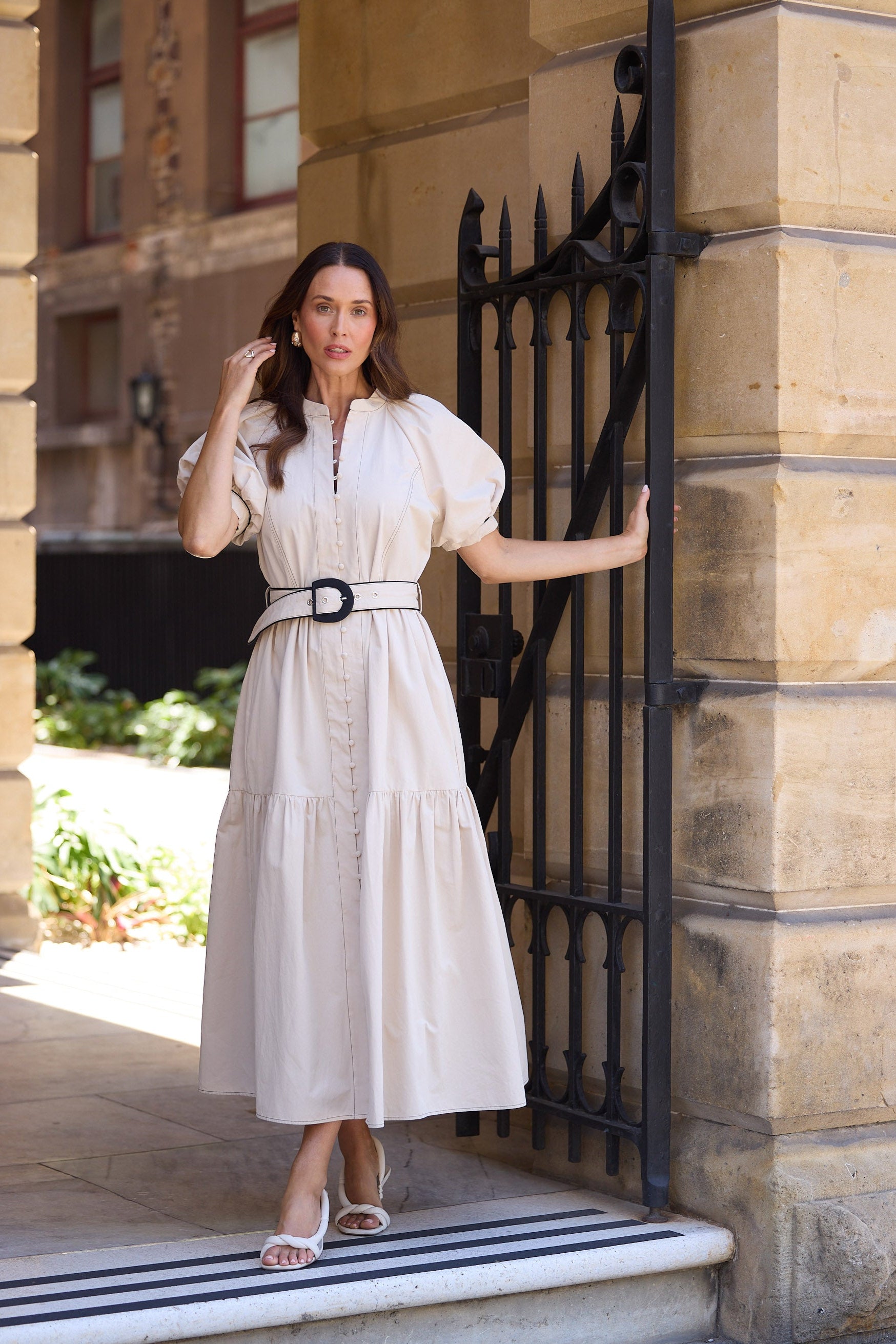 Woman in a white dress standing in front of a black iron gate on a sunny day.