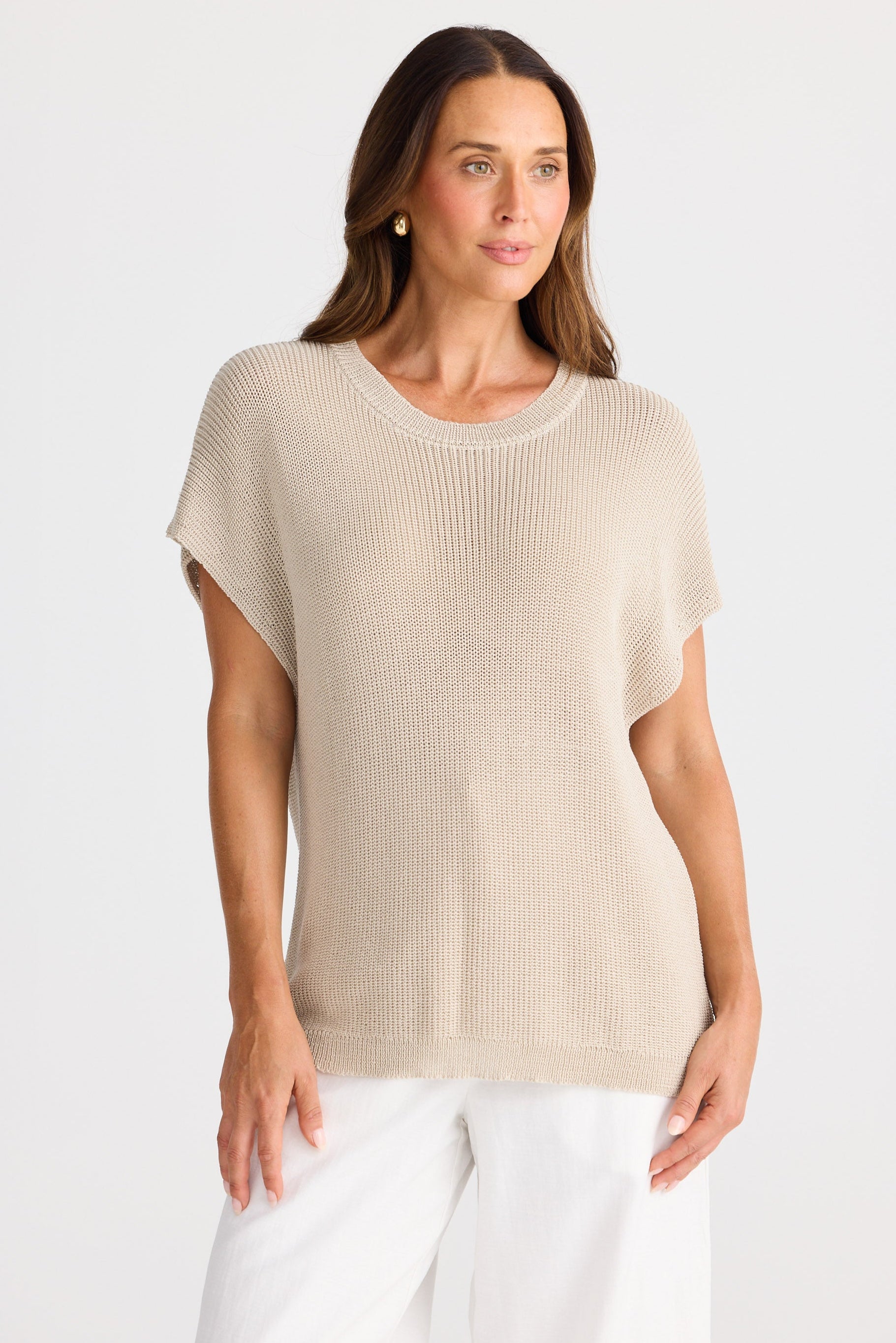 Woman wearing a beige knit top and white pants on a light gray background