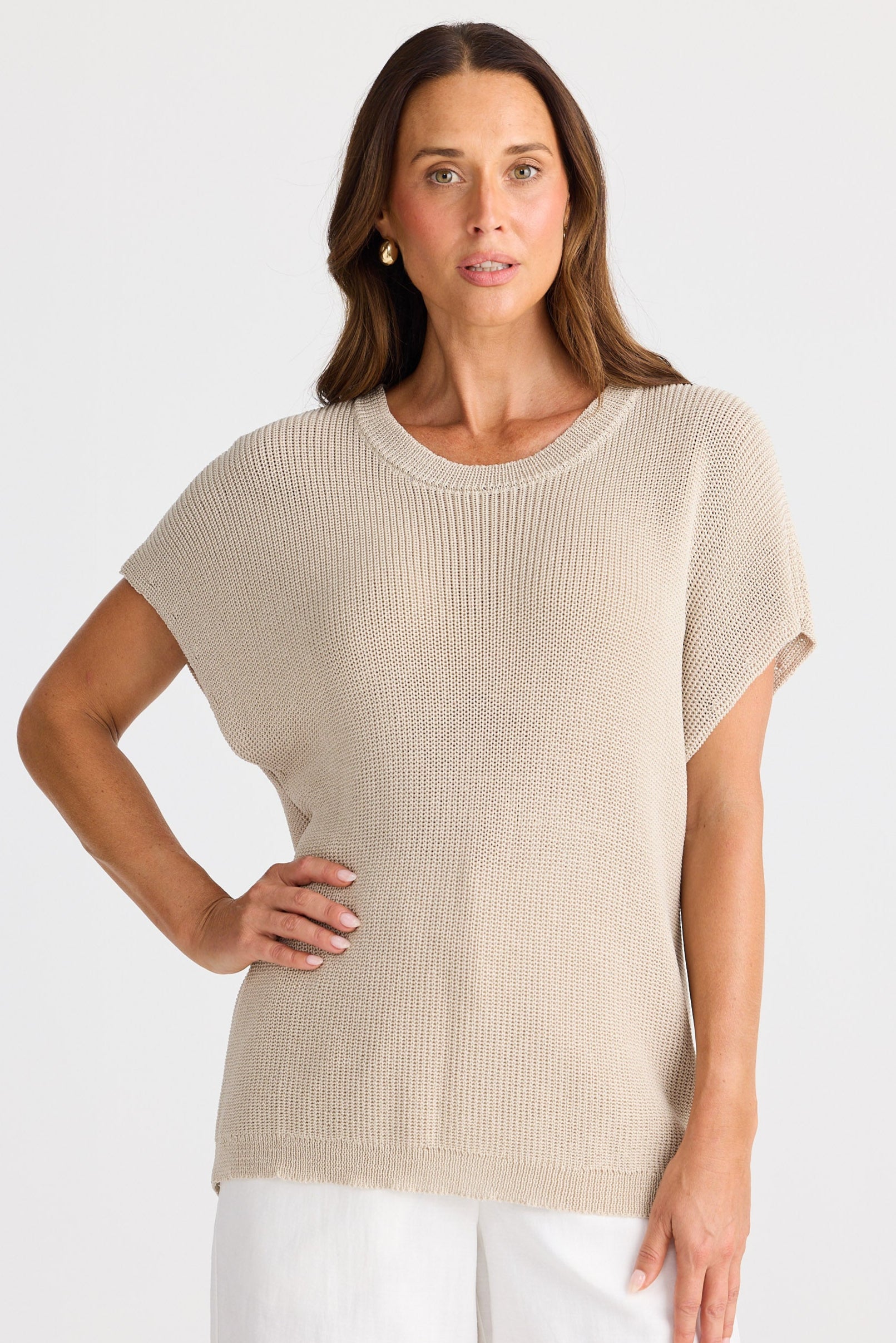 Woman wearing a beige knit top and white pants on a plain background