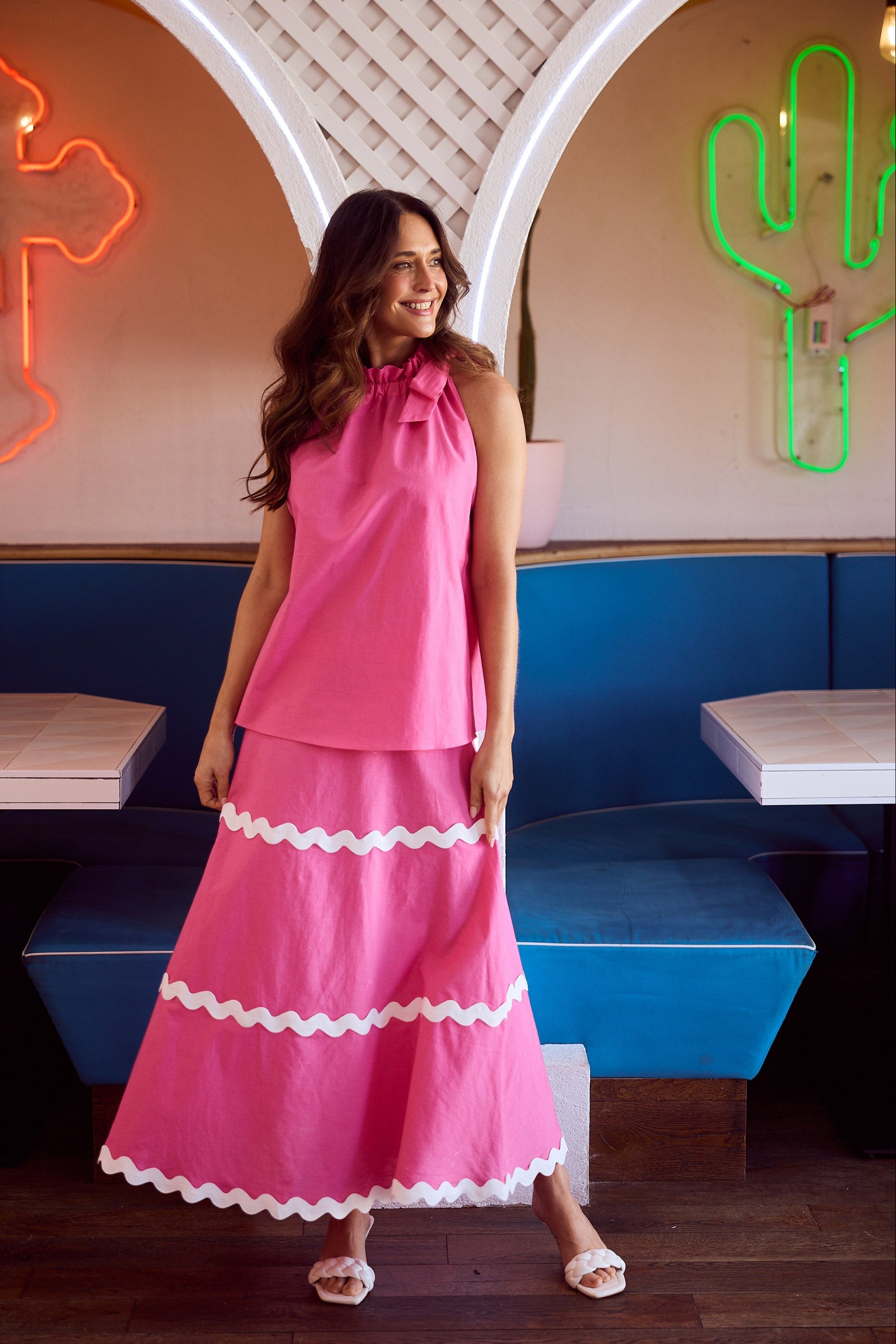 Woman in a pink top and skirt standing in a retro-style diner with neon signs.