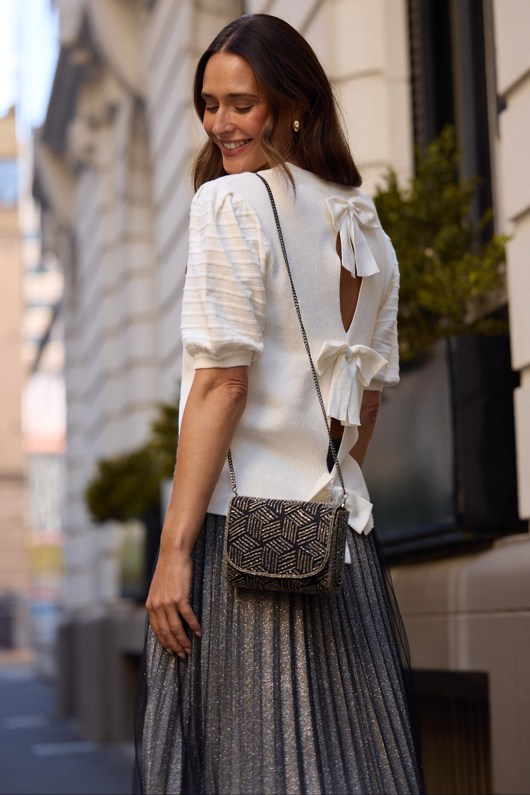 Woman wearing a white knit top with bow details and a pleated skirt, standing on a city street.