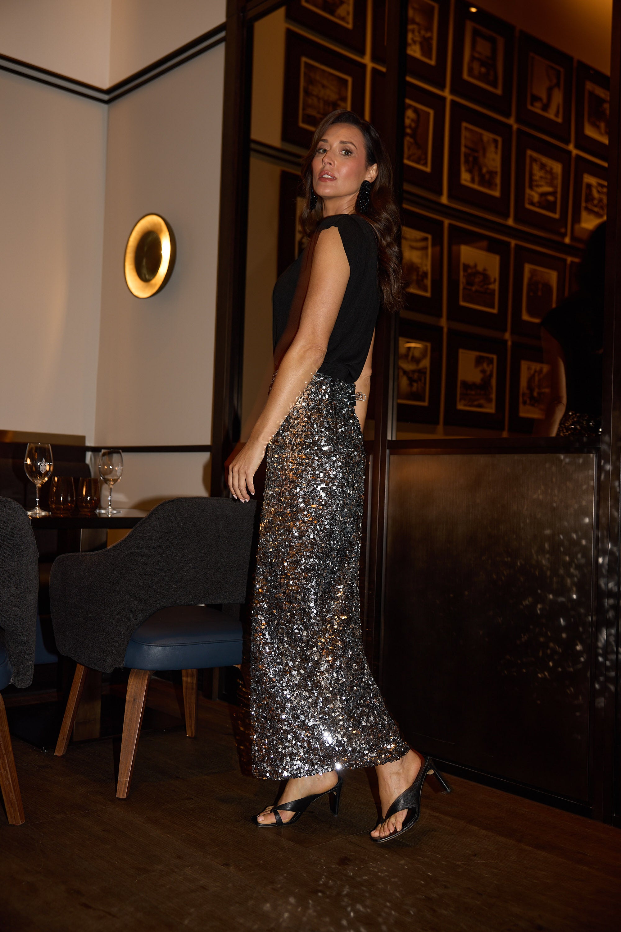 Woman in a black top and silver sequin skirt standing in an elegant room with framed pictures on the wall.