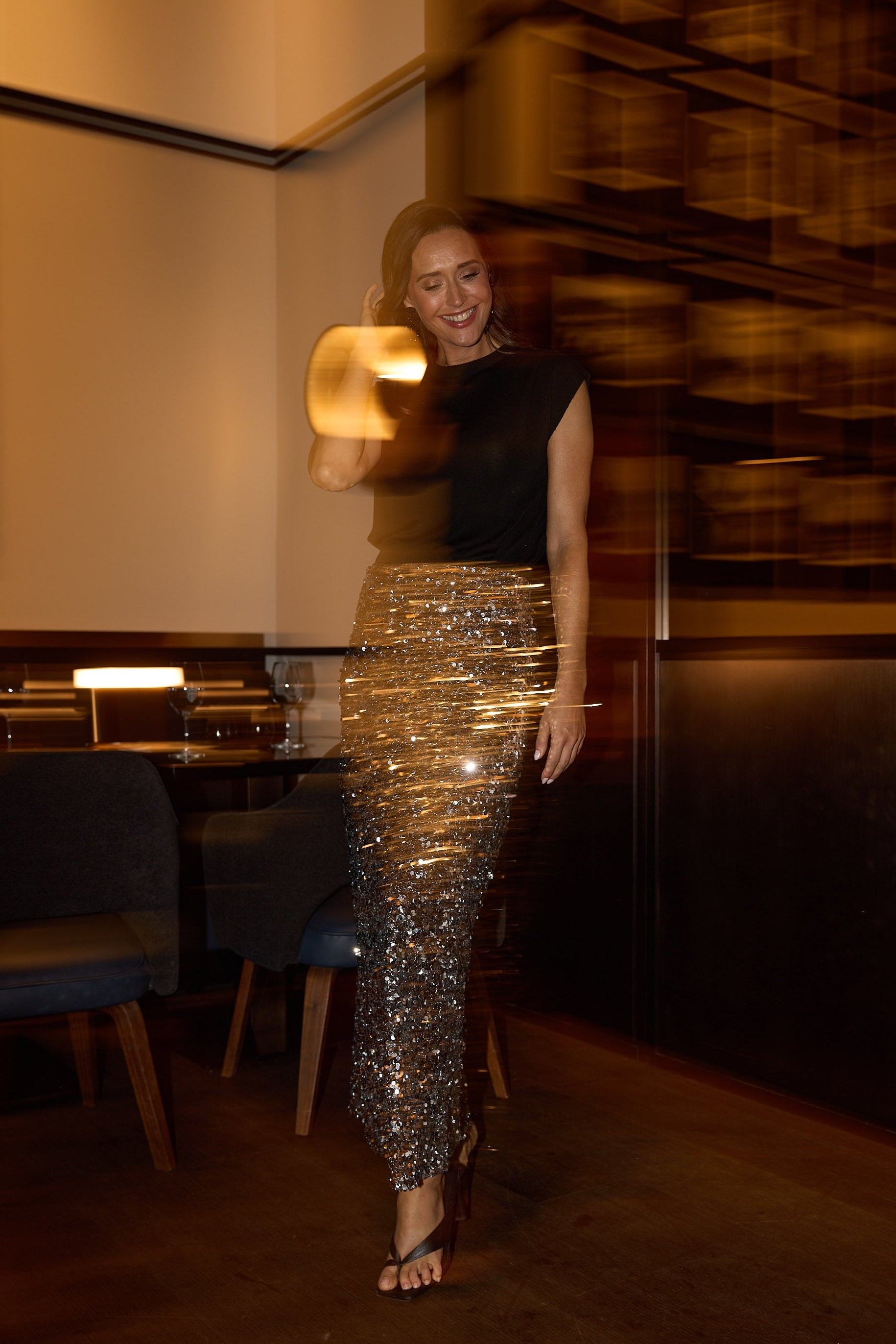 Woman in a black top and gold sequin skirt standing in a dimly lit room with wooden shelves.