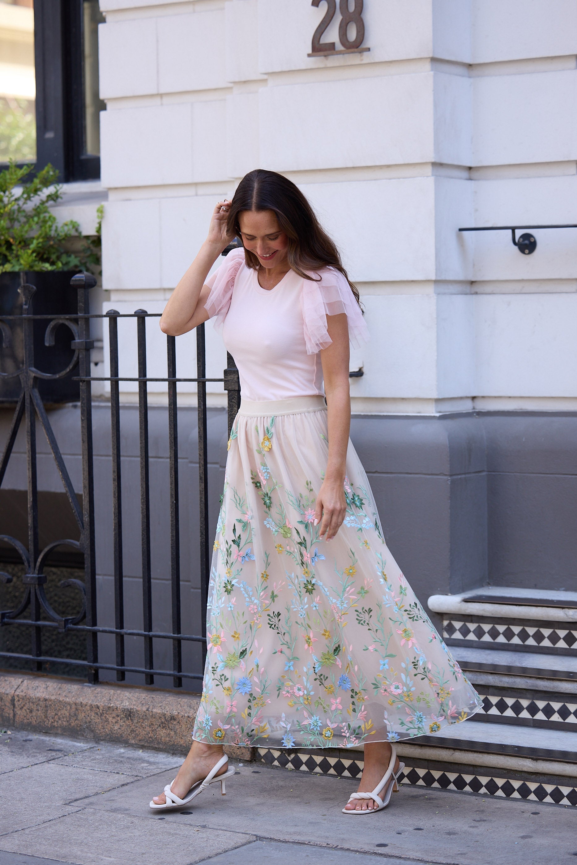 Woman in a light pink top and floral skirt standing on a sidewalk with a building in the background.