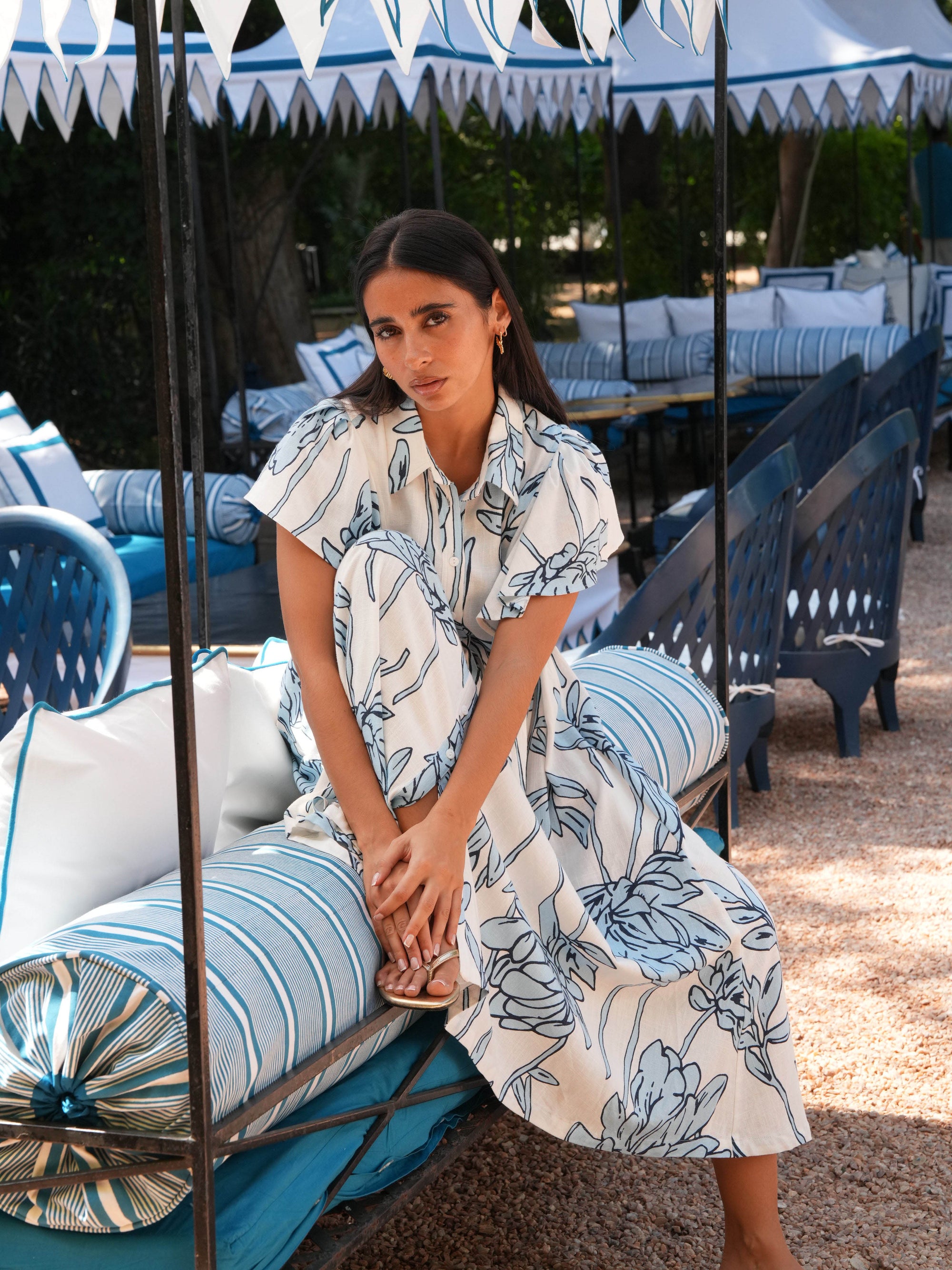 Woman in a floral dress sitting on a lounge chair under a canopy with striped chairs and tables in the background.