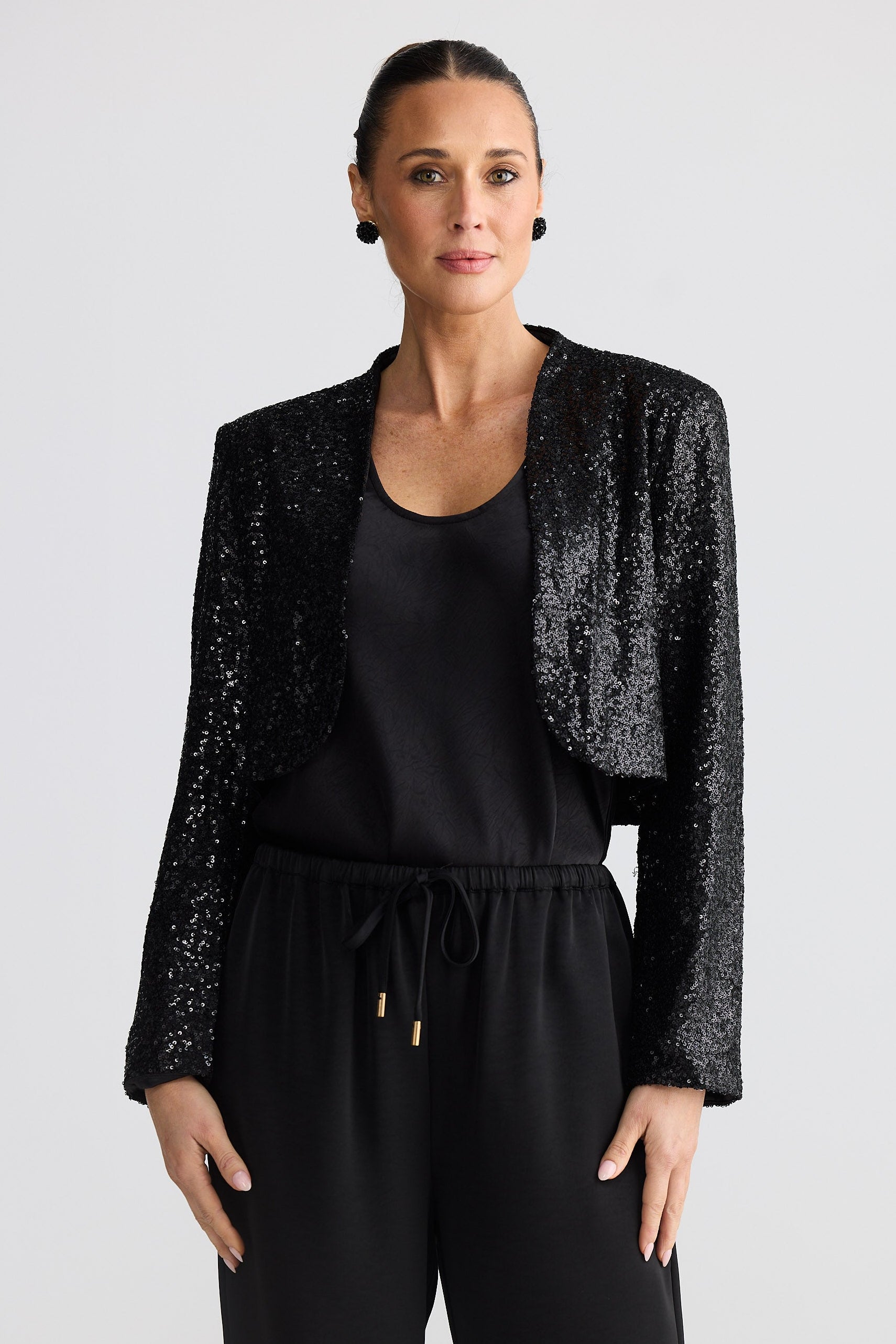 Woman wearing a black sequin jacket over a black top and pants on a light gray background