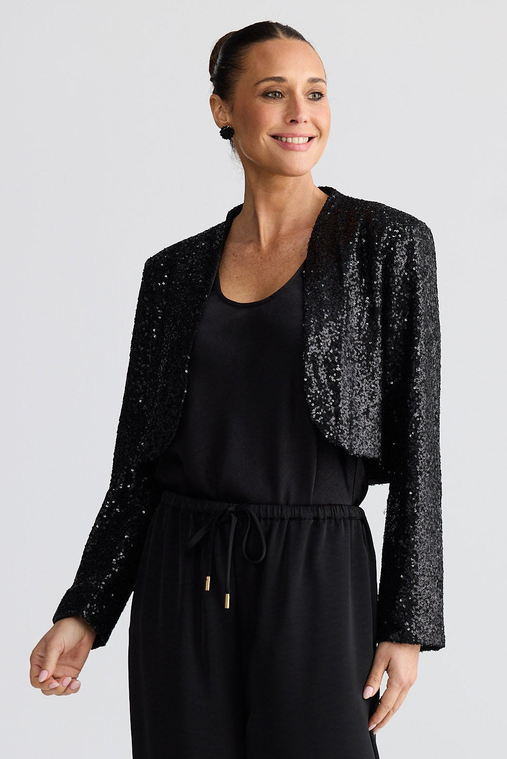 Woman wearing a black top and pants with a sparkly black cardigan on a light gray background
