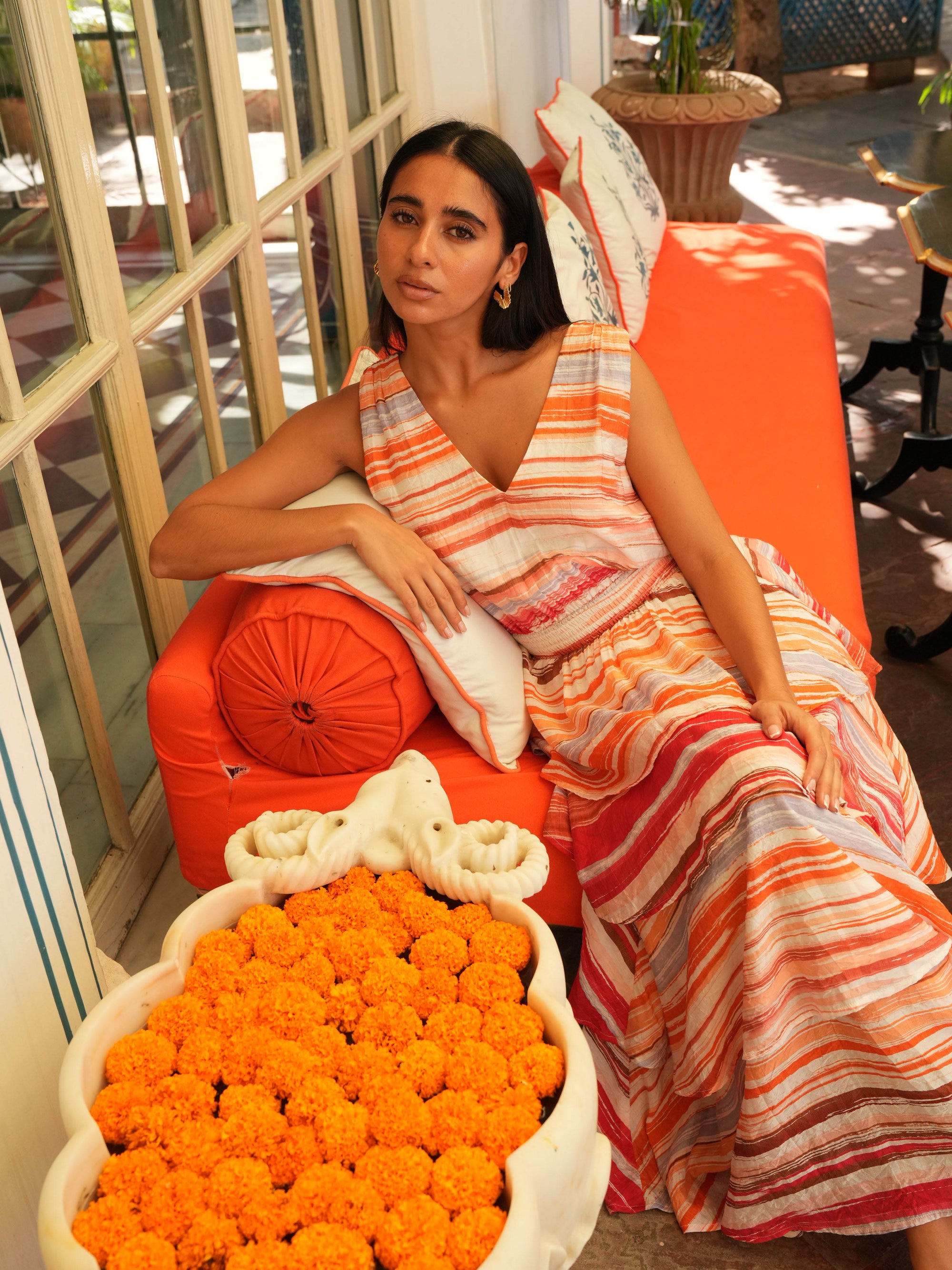 Woman in a colorful dress sitting on an orange chair with a large bowl of orange flowers in front of her.
