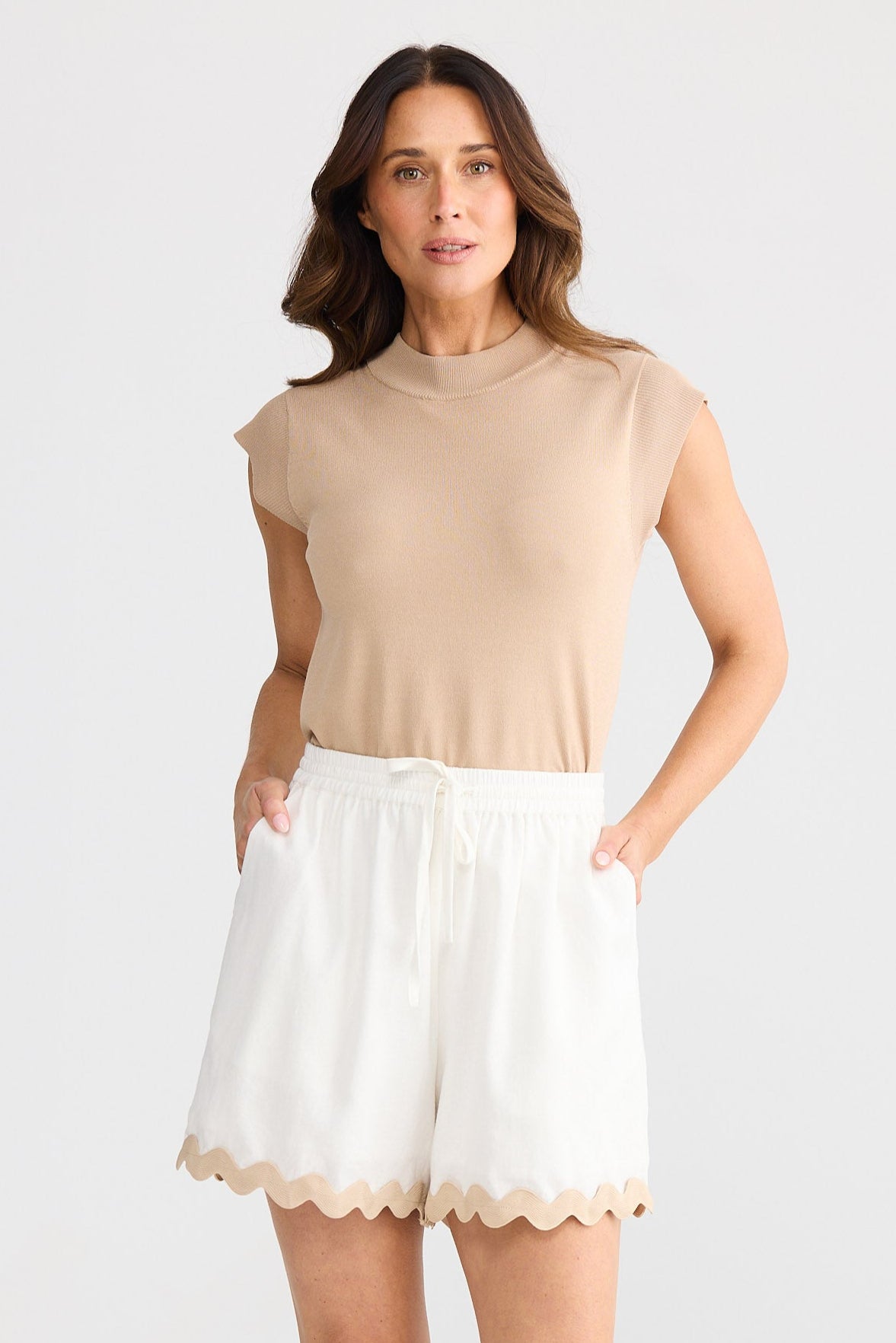 Woman wearing a beige sleeveless top and white shorts with ric rac trim on a white background