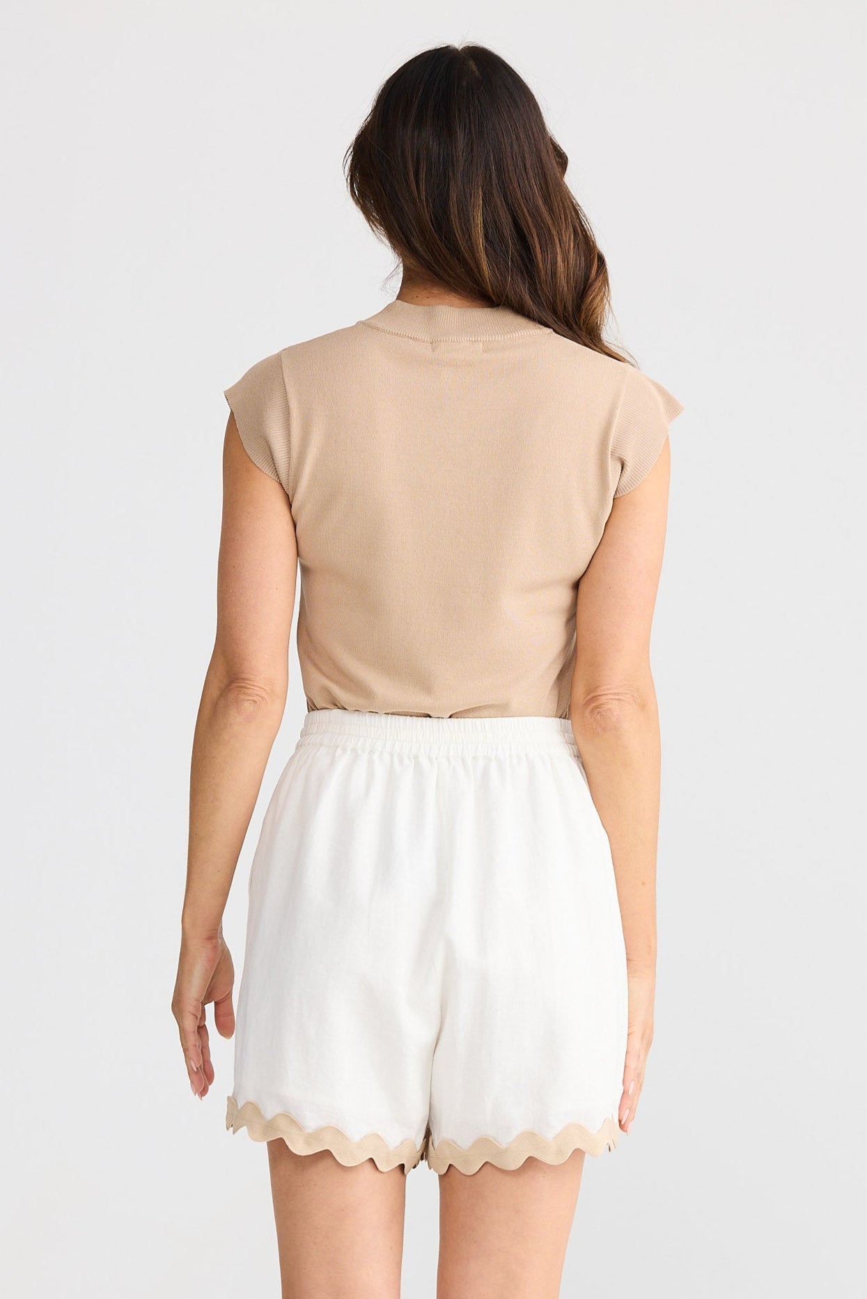 Woman wearing a beige sleeveless top and white shorts with ric rac trim on a plain background