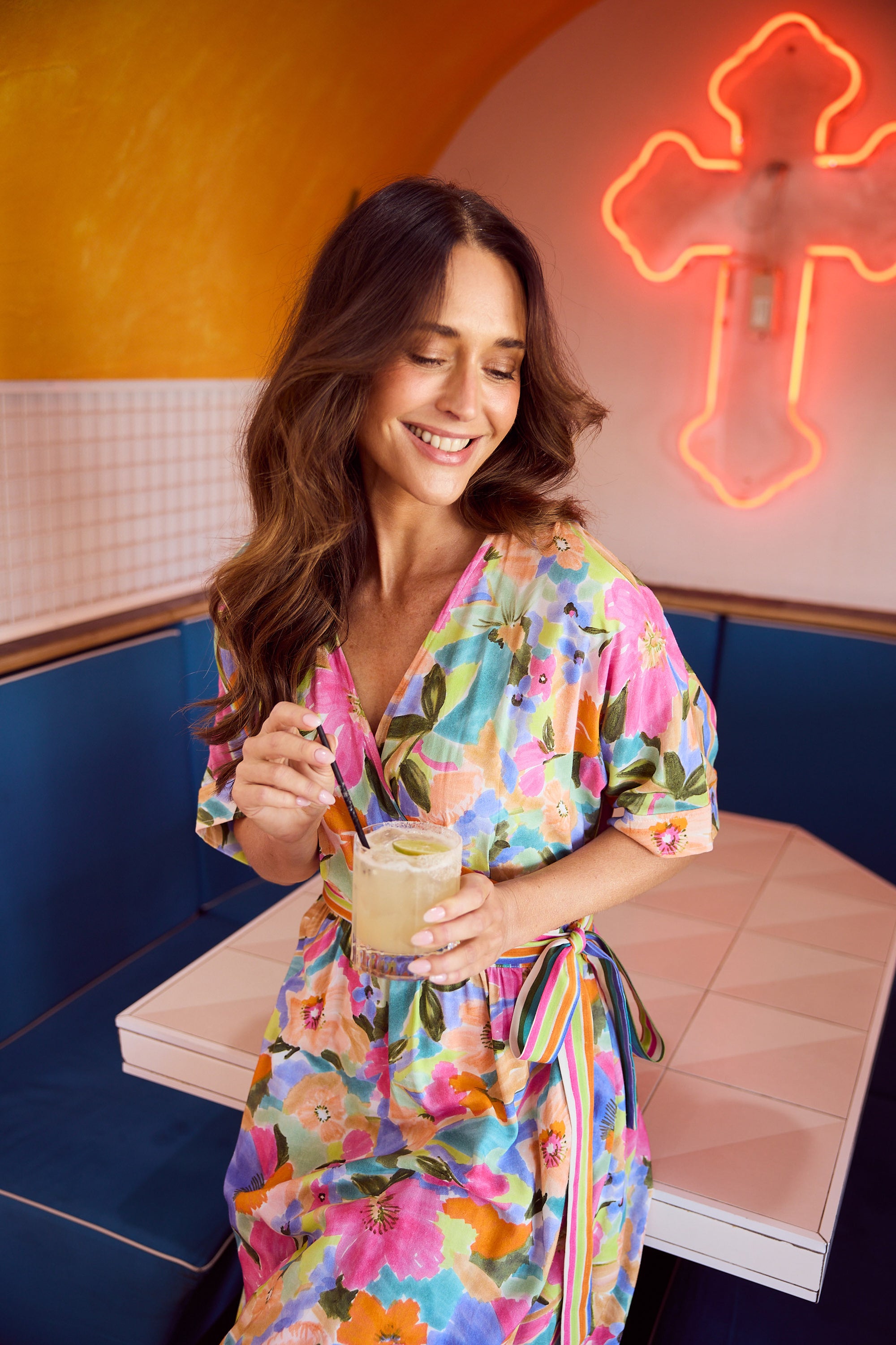 Woman in a colorful dress holding a drink in a retro setting with a neon sign.