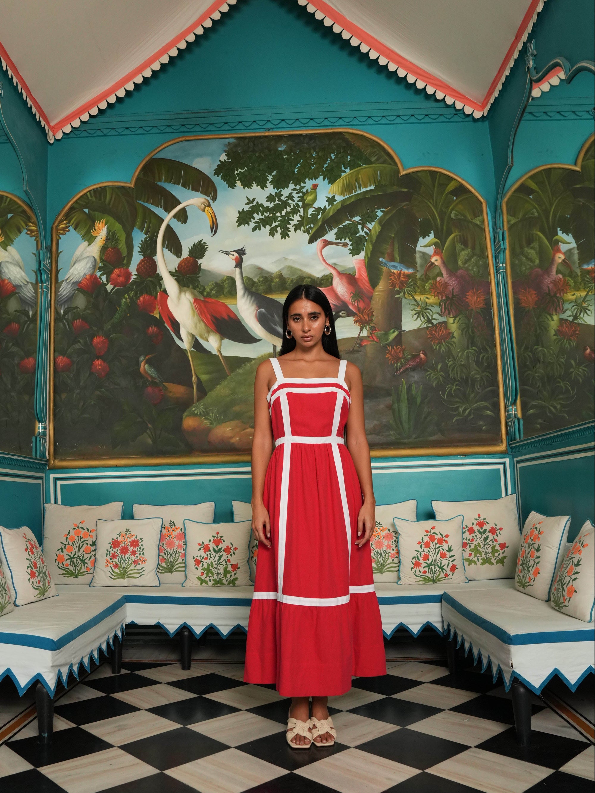 Woman in a red dress standing in a decorated room with floral cushions and a checkered floor.