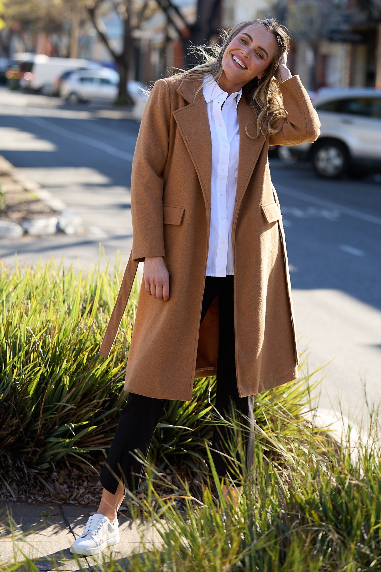 Woman in a tan coat standing on a street with cars and buildings in the background