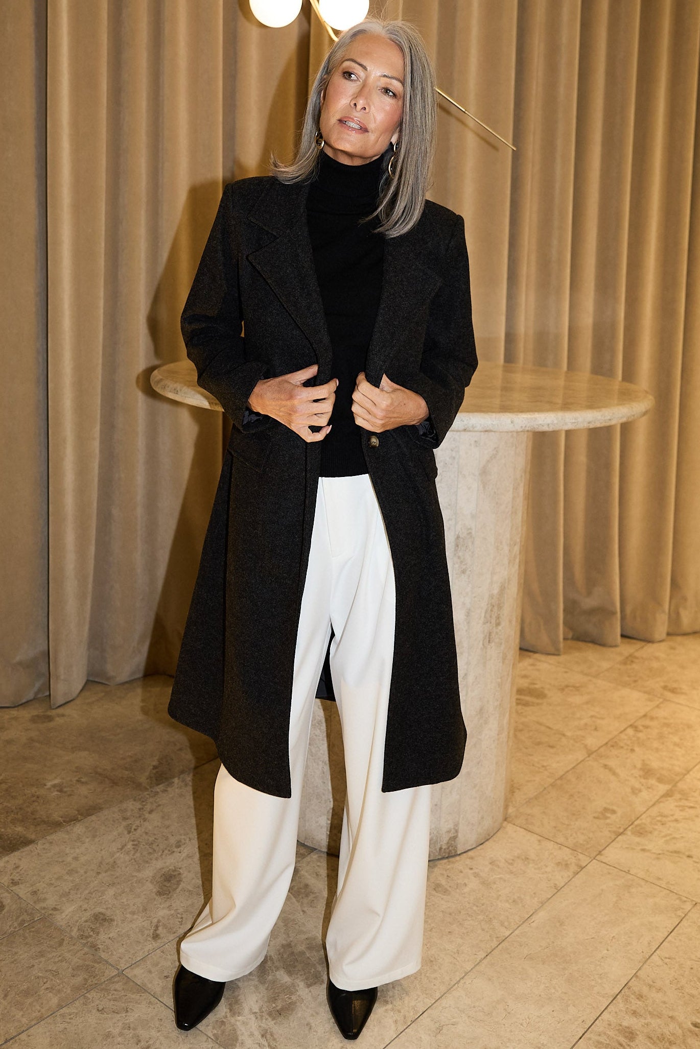 Person wearing a black coat and white pants standing in a room with beige curtains and a decorative light fixture.