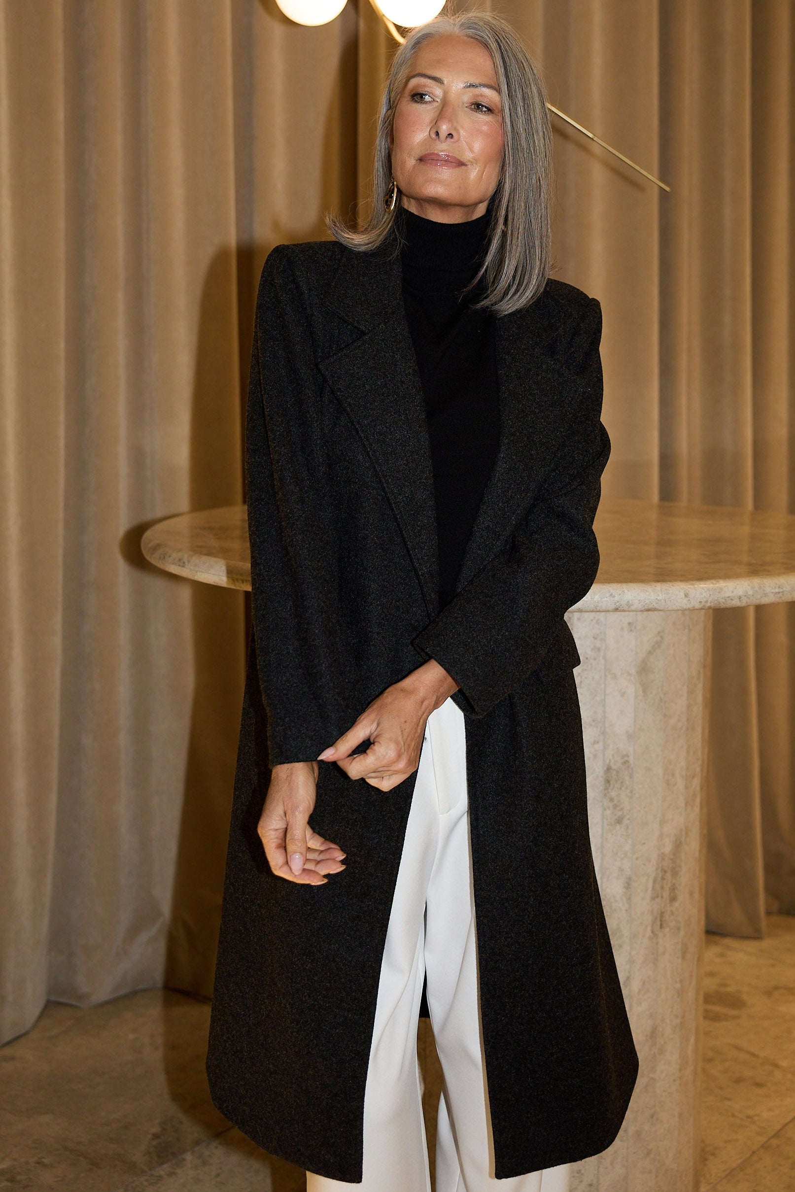 Woman in a black coat standing in a room with beige curtains and a round table.