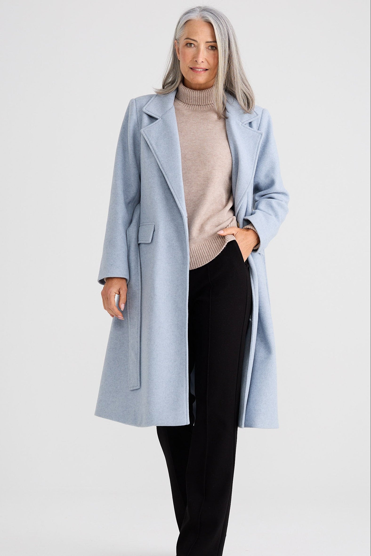 Woman wearing a light blue coat over a beige sweater and black pants on a white background