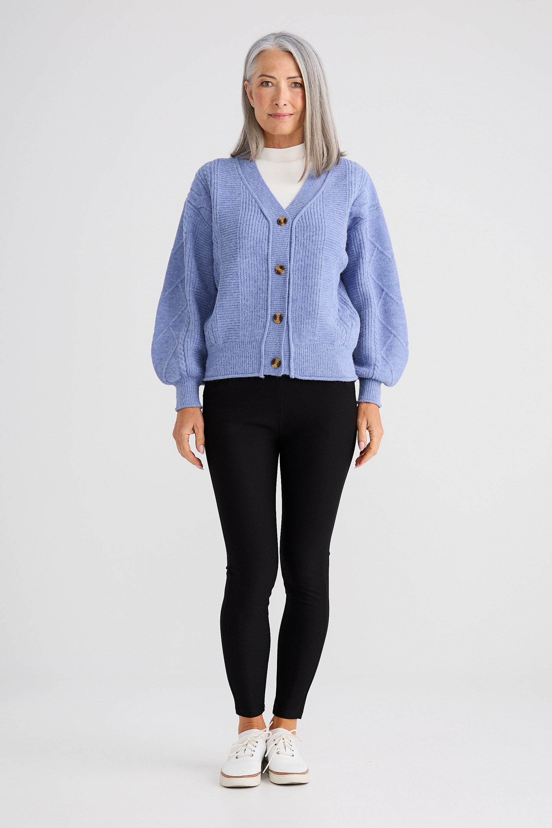 Woman wearing a blue cardigan over a white shirt, black pants, and white sneakers on a white background