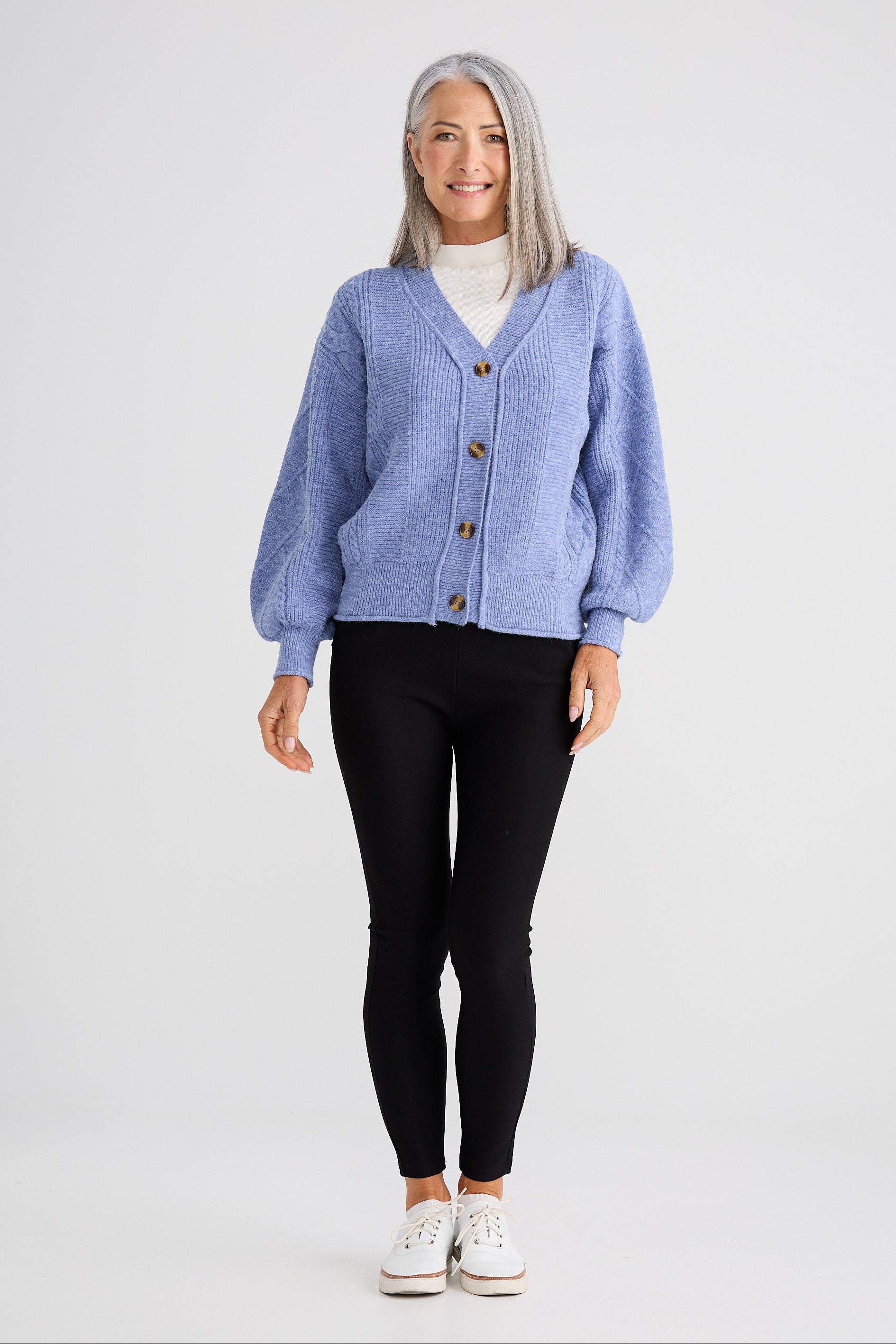 Woman wearing a light blue cardigan over a white shirt, black pants, and white sneakers on a white background.