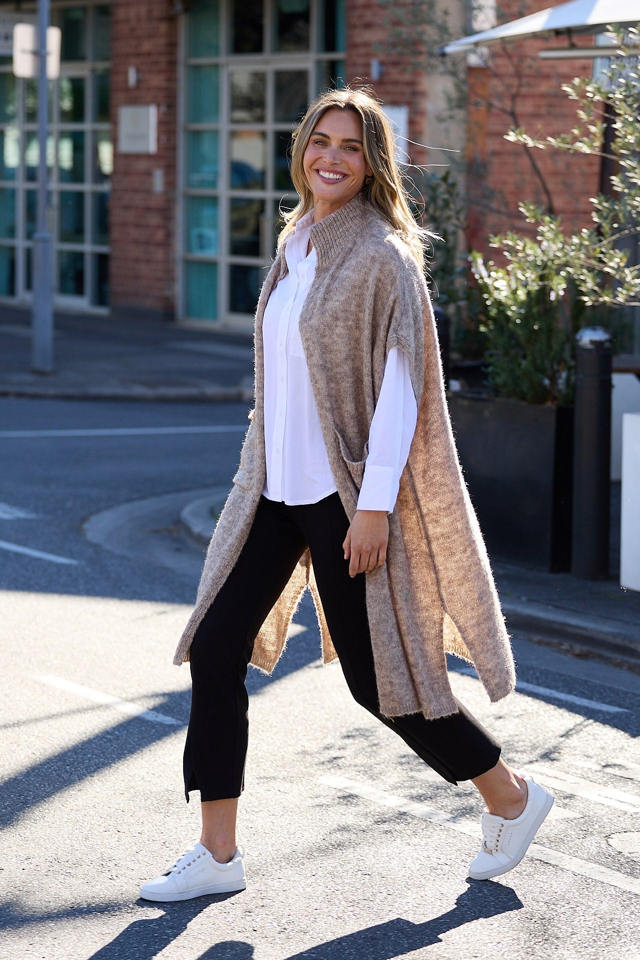 Woman walking on a street wearing a beige cardigan, white shirt, black pants, and white shoes.