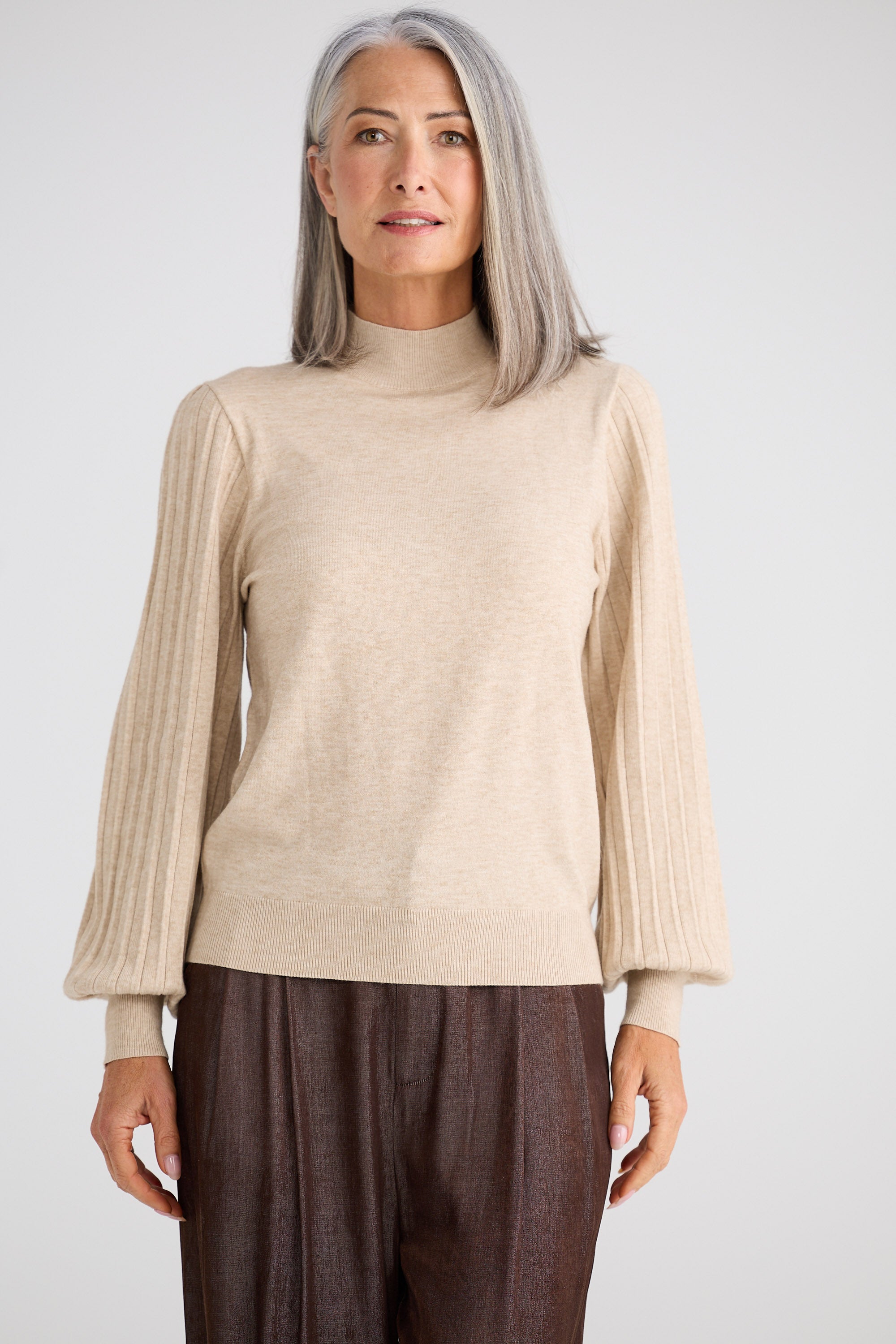Woman wearing a beige sweater and brown pants against a white background