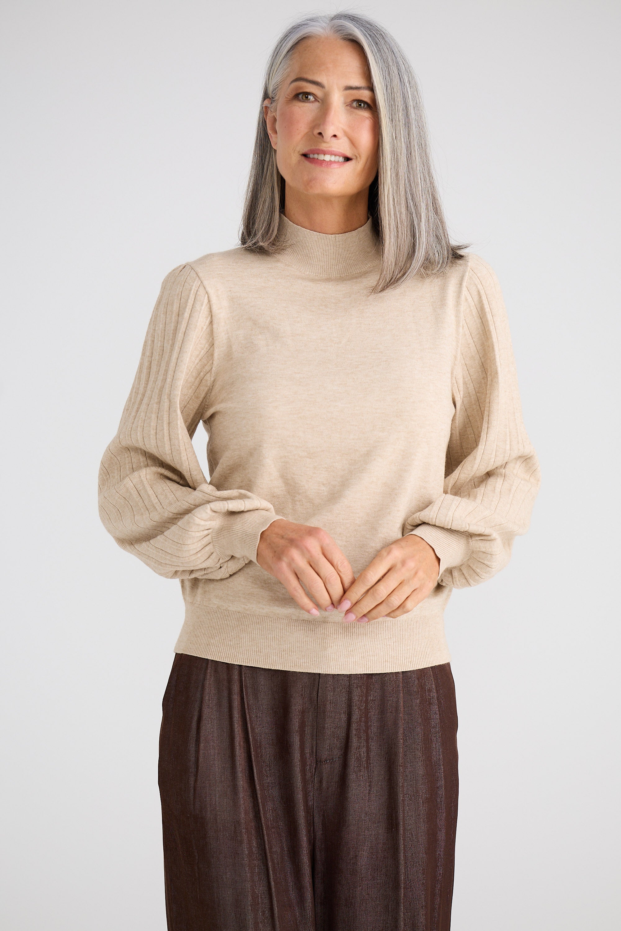 Woman wearing a beige sweater and brown pants against a white background