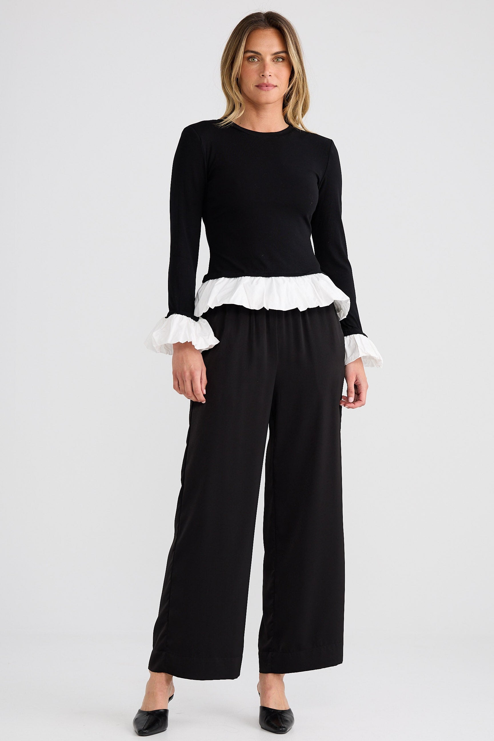 Woman wearing a black top with white ruffle details and black pants on a white background