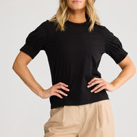 Woman wearing a black top and beige pants against a plain background
