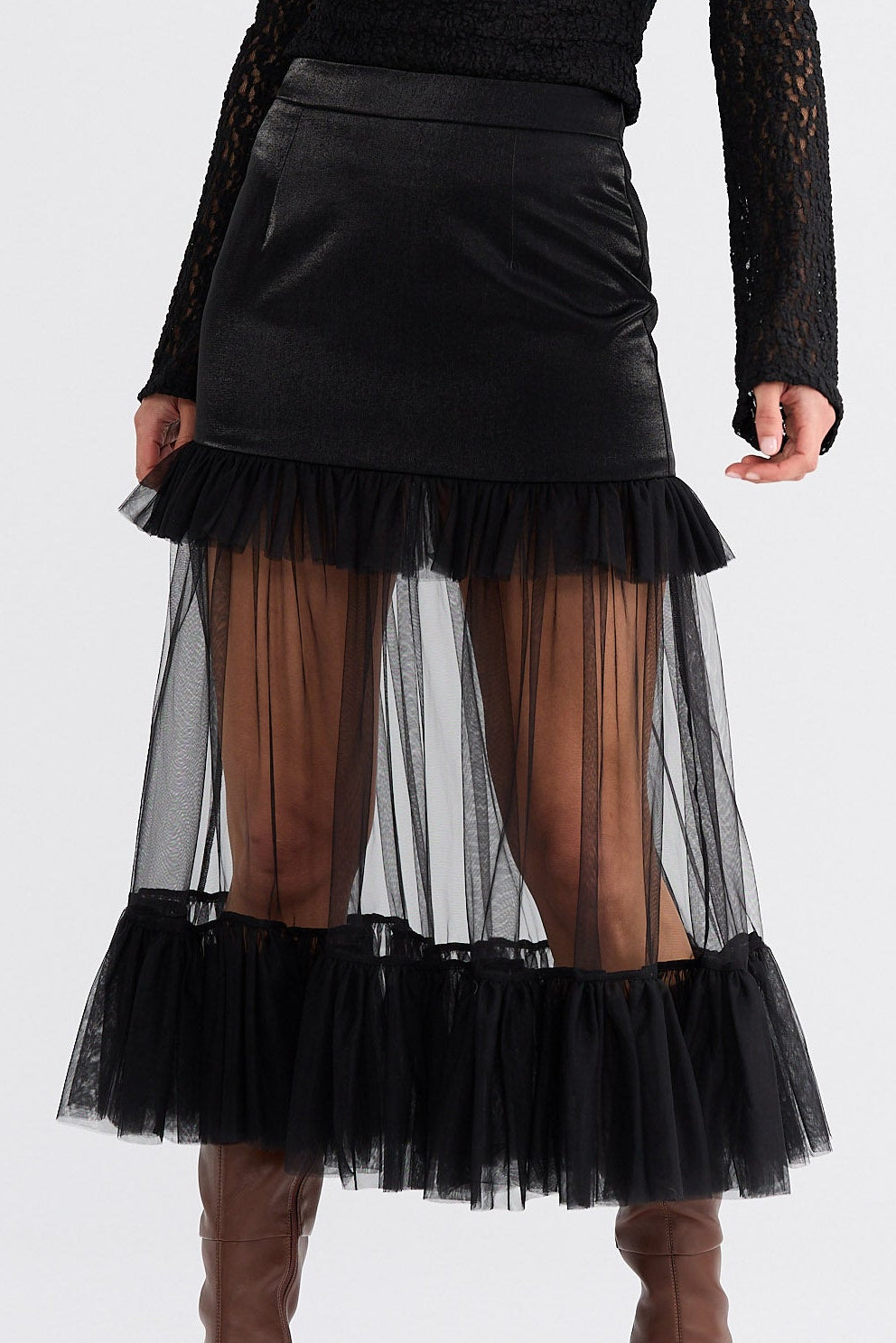 Woman wearing a black turtleneck with sheer sleeves, black skirt with sheer bottom half, and brown boots on a white background.