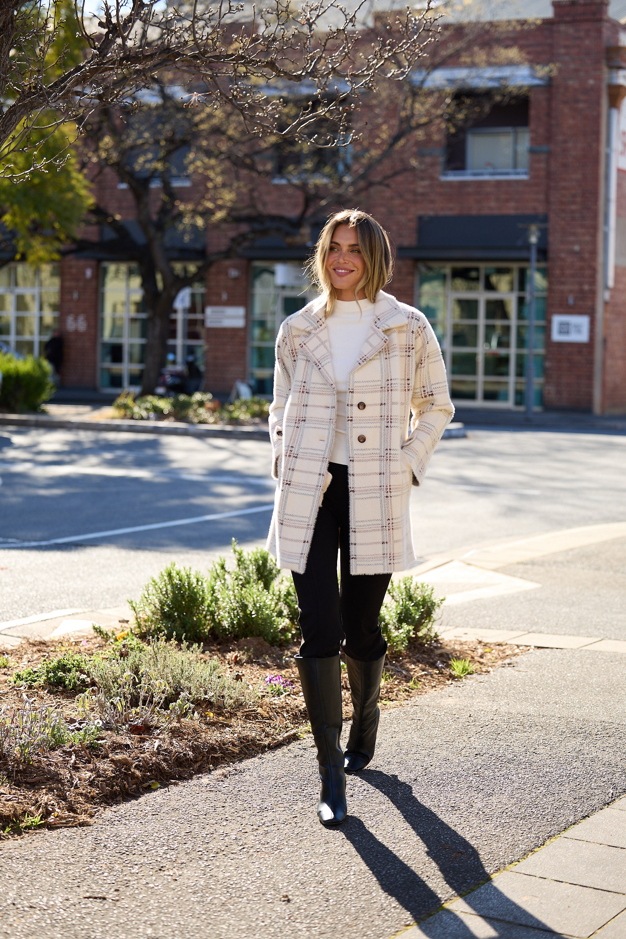 Woman walking on a sidewalk wearing a plaid coat and dark pants.
