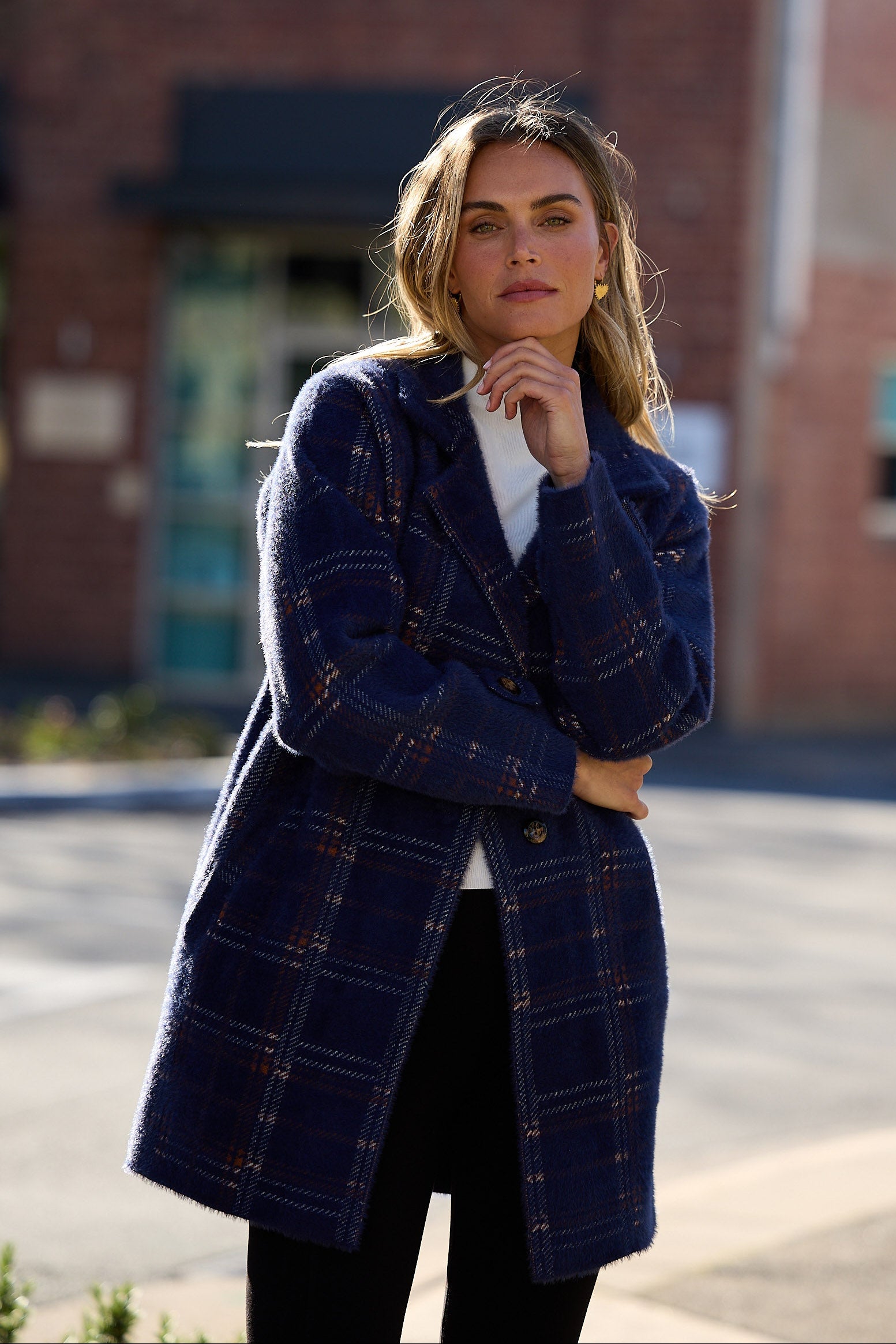 Woman wearing a blue plaid coat standing on a street with a brick building in the background