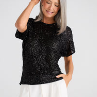 Woman wearing a black sequin top and white pants against a plain background