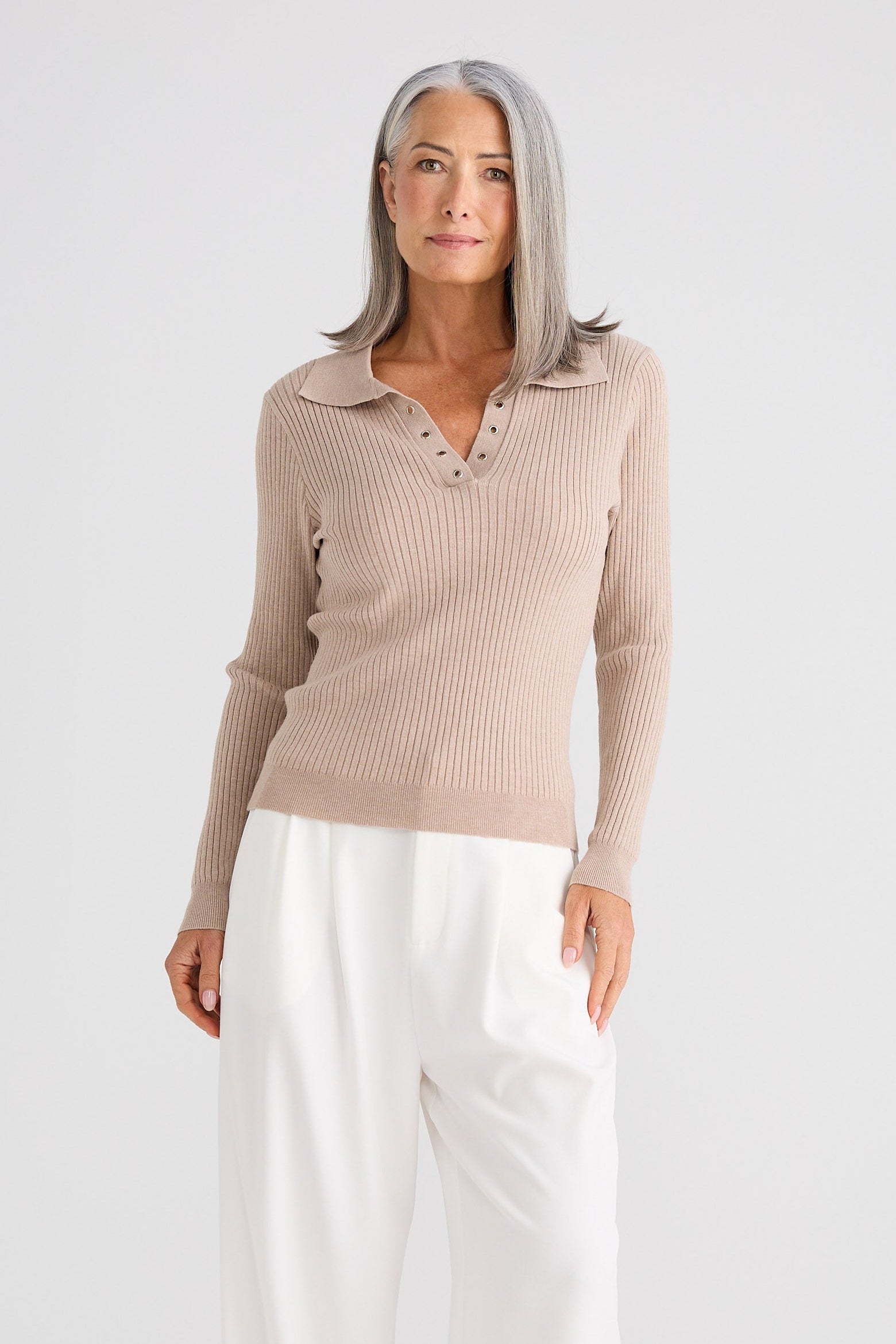 Woman wearing a beige ribbed sweater and white pants on a white background