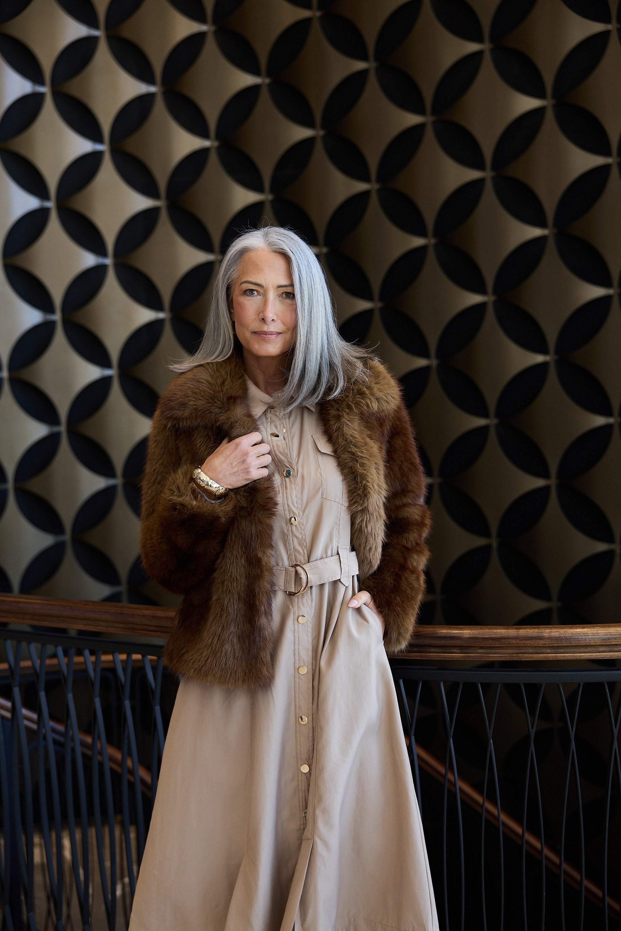Woman wearing a fur coat and beige dress against a patterned wall.